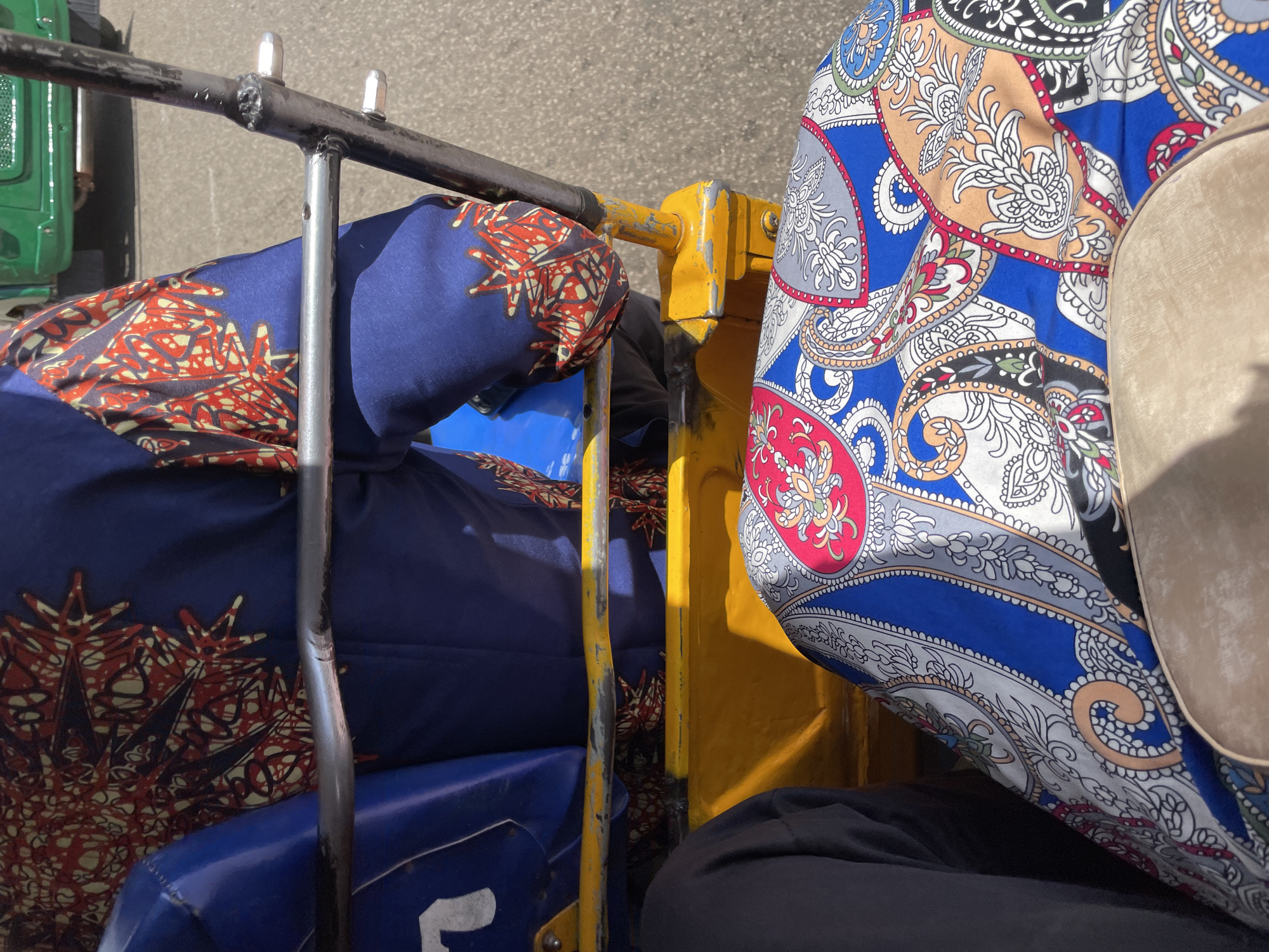 The image depicts a close-up, high-angle view from inside a motorized rickshaw, commonly known as an "Okada" or "Keke Napep," in Jos, Nigeria. The foreground is dominated by a seat covered in a vibrant, colorful fabric with a paisley-inspired pattern featuring blue, pink, white, grey, and orange elements. To the left, a person's dark gray trousers are visible, suggesting they are sitting next to the viewer.
Behind the front seat, a person wearing a blue garment with a striking red, black, and yellow patterned print is seated. This garment appears to be a long-sleeved top or jacket with intricate designs on the sleeves. A metal bar, part of the rickshaw's frame, runs horizontally across the middle of the image, obscuring a portion of the person's torso and the yellow painted body of the rickshaw. A blue plastic seat is partially visible behind the metal bar.
In the background, a green vehicle, likely a truck or bus, is seen on a paved road, indicating an urban or semi-urban environment. The sunlight suggests it is daytime, and the asphalt road is visible. The overall impression is a snapshot of daily life and transportation in Jos, Nigeria, focusing on the distinct patterns of the clothing and the rickshaw's interior.