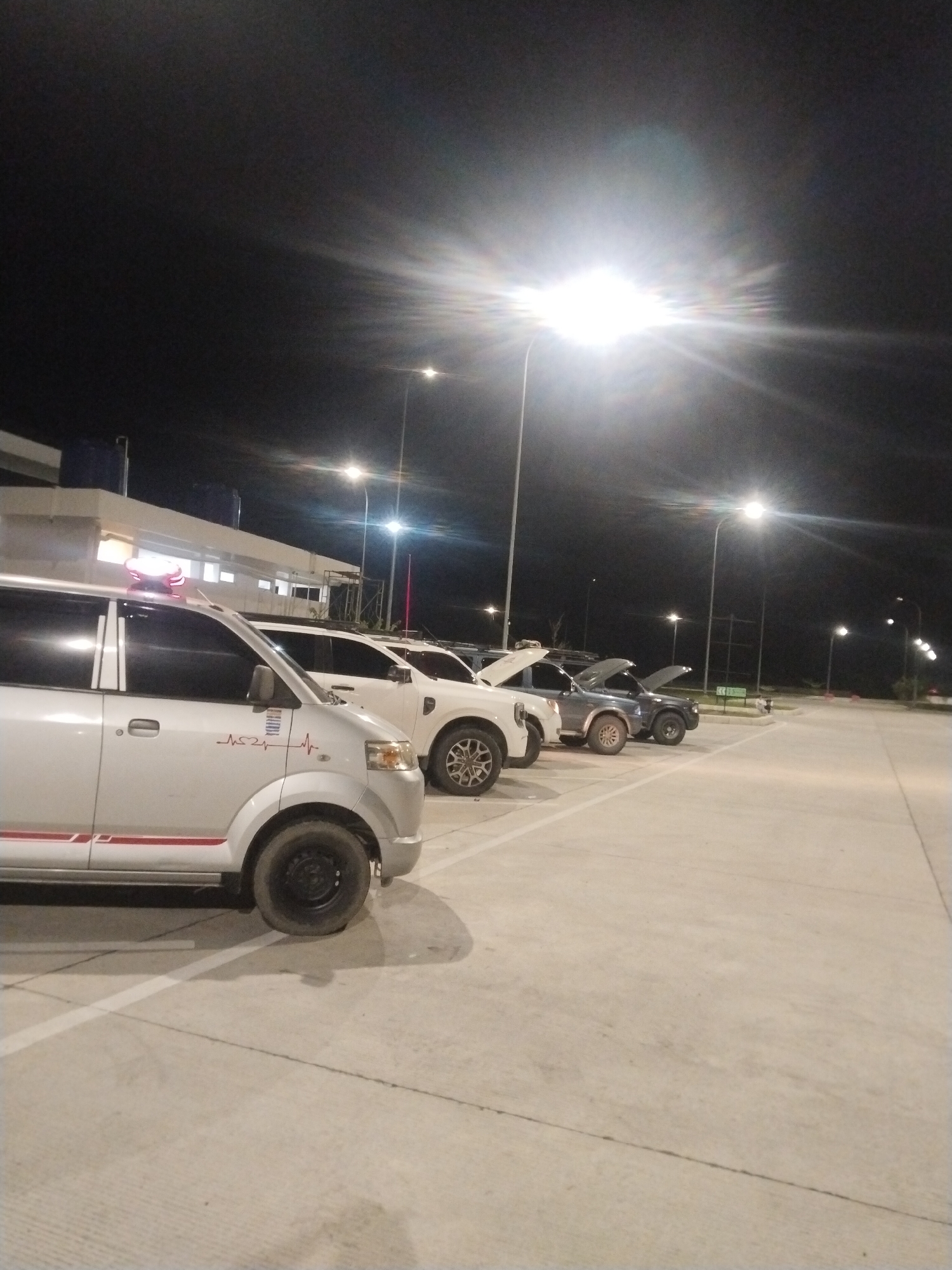 The image shows a row of vehicles parked in a brightly lit parking lot at night. The location is in the city of Keureuweungkrueng, Indonesia. The first vehicle in the row is a light-colored van with a red stripe and a graphic resembling a heart rate monitor. Behind it is a white pickup truck and two other vehicles with their hoods open. There is a building with a flat roof in the background and several streetlights illuminating the parking lot and the surrounding area. The sky is dark, and the parking lot is large and mostly empty.