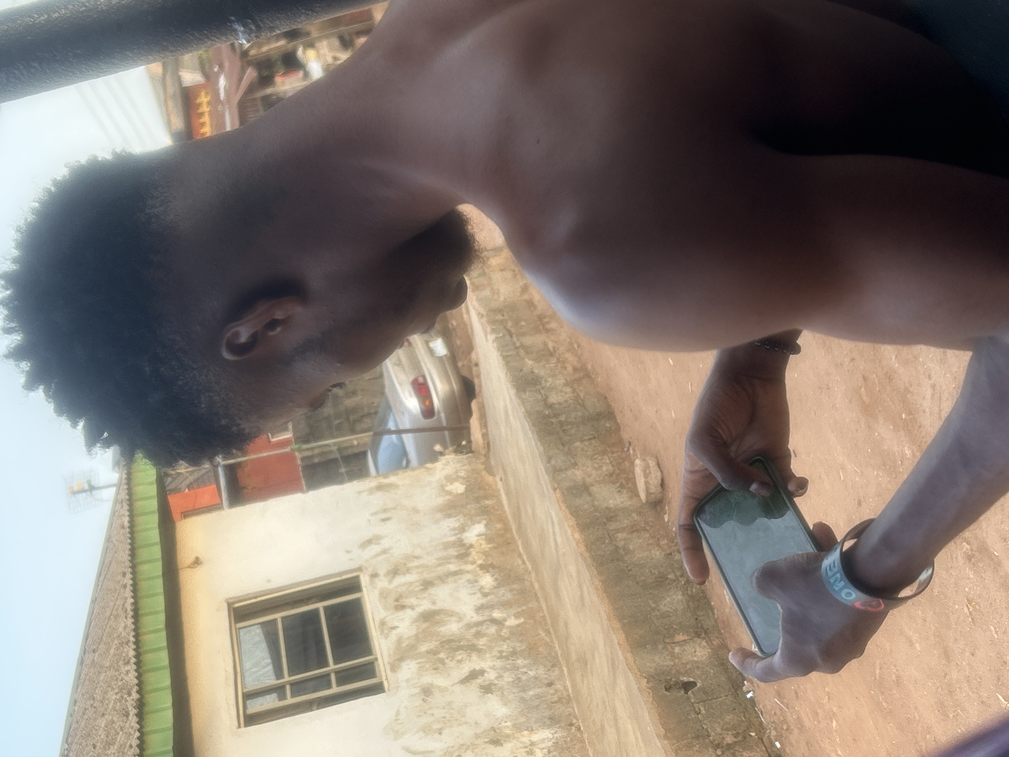 An outdoor, slightly askew shot captures a shirtless man from a side-back perspective, standing in what appears to be a residential or semi-urban area. He has short, dark, curly hair and a light beard or stubble. The man is holding a smartphone horizontally with both hands, positioned as if taking a photo or video, or possibly interacting with the screen. A dark metal bar or railing is visible in the upper foreground.
On his right wrist, he wears a light-colored bracelet with the visible text "ONE C". The background features various buildings, including a light-colored structure with a multi-paned window, and another building with green corrugated siding and what appears to be an antenna or similar structure on its roof. A silver car is partially visible behind the buildings. The ground beneath him is a light, dusty brown. The lighting suggests it is daytime with clear weather.
