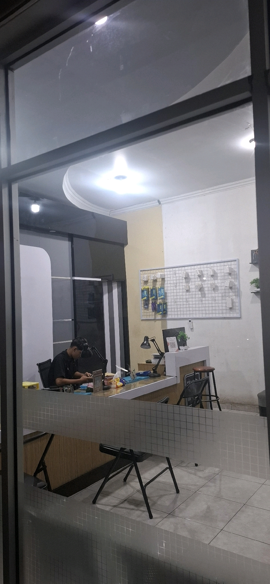 The image shows the interior of what appears to be a small repair shop or office, viewed through a glass window. A man wearing a dark shirt and headphones is seated at a counter, engaged with something on his hands, possibly a repair. The counter is equipped with a desk lamp, various tools, and what seems to be electronic components. In the background, a grid display with small items is mounted on the wall, and a monitor and a small plant are visible on the counter. The overall lighting suggests it is nighttime, with artificial lights illuminating the space. The presence of a counter and display suggests a retail or service-oriented establishment. The location is identified as Surakarta, Indonesia.