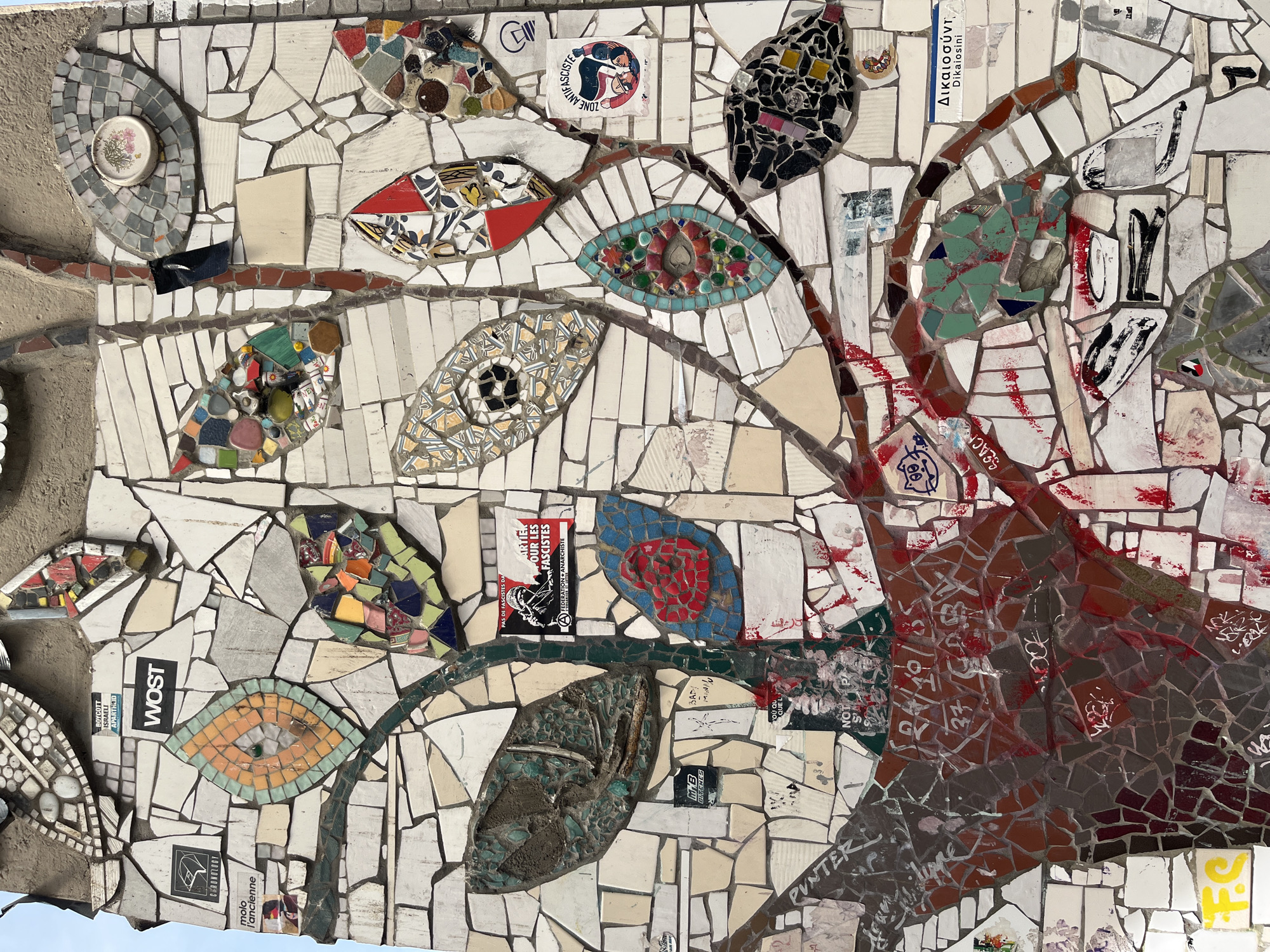 The image displays a section of a mosaic art installation covering a wall, possibly outdoors given the lighting and sky visible at the top. The mosaic is composed of numerous irregularly shaped pieces of tile, ceramic, and other materials, forming various patterns and images. Prominent motifs include stylized leaves in green, orange, and blue, as well as abstract geometric designs. Several small stickers and posters are integrated into the mosaic, some with text and graphics. One sticker features an anarchist symbol and the text "PAS DE FA SCI ST ES" and "FEDERATION ANARCHISTE". Another sticker contains Greek text that translates to "Justice". There are also crude drawings and graffiti applied over parts of the mosaic, including what appears to be dates and numbers like "24/10/85" and "27/10/83". The color palette is diverse, featuring white, cream, grey, green, red, blue, orange, and black. The overall impression is one of a vibrant, layered, and potentially political street art piece. This artwork is located in the Belleville neighborhood of Paris, France, and is part of a larger mosaic project found on a building at 26 Rue Dénoyez.