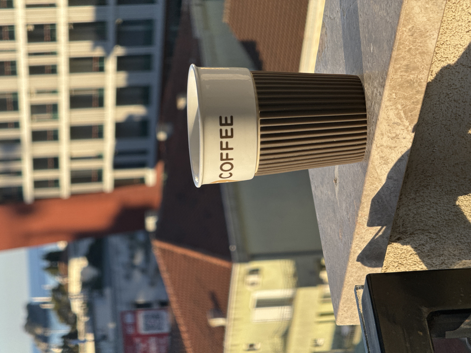 A close-up shot focuses on a coffee cup with the word "COFFEE" printed on it, placed on a ledge in the foreground. The cup is white with a brown ribbed sleeve. In the background, a blurred cityscape is visible, featuring buildings with red roofs and a larger, modern building with many windows. The scene is bathed in the warm light of what appears to be late afternoon or early morning. The image is taken from a high vantage point, suggesting a view from a balcony or rooftop overlooking the city of Budva, Montenegro. The sea is faintly visible in the distance. There are no people or animals present in the image.