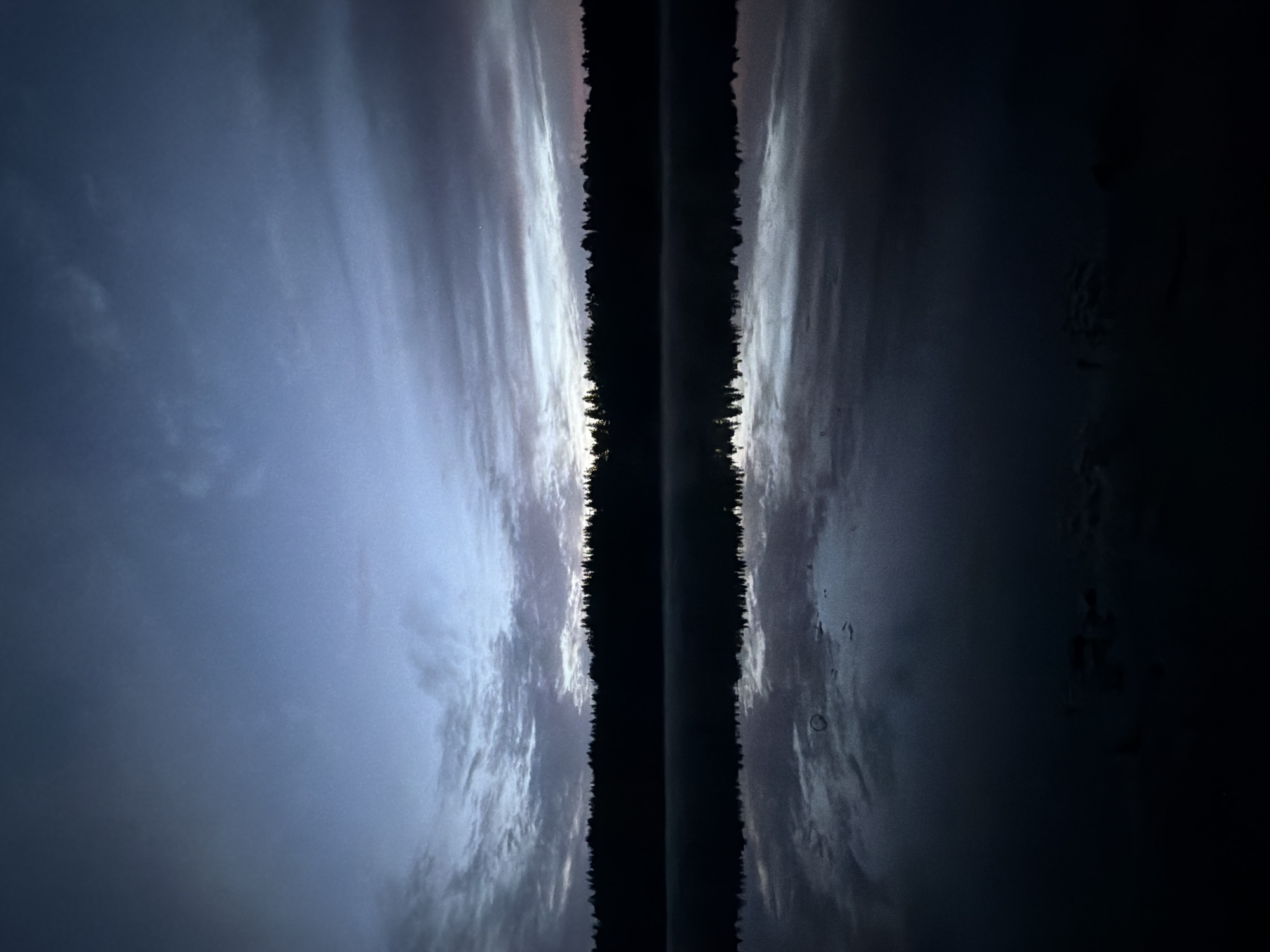 The image captures a twilight scene over a body of water, likely a lake, in Solov'evo, Russia. A dense line of silhouetted trees forms a dark horizon across the middle of the frame, separating the sky from the water. The sky above is a gradient of deep blues and grays, with scattered clouds exhibiting subtle pink and orange hues from the setting or rising sun. The water is calm and reflective, mirroring the sky and the treeline. In the lower portion of the water's reflection, faint lighter areas suggest movement or possibly very shallow water. The overall atmosphere is serene and tranquil.