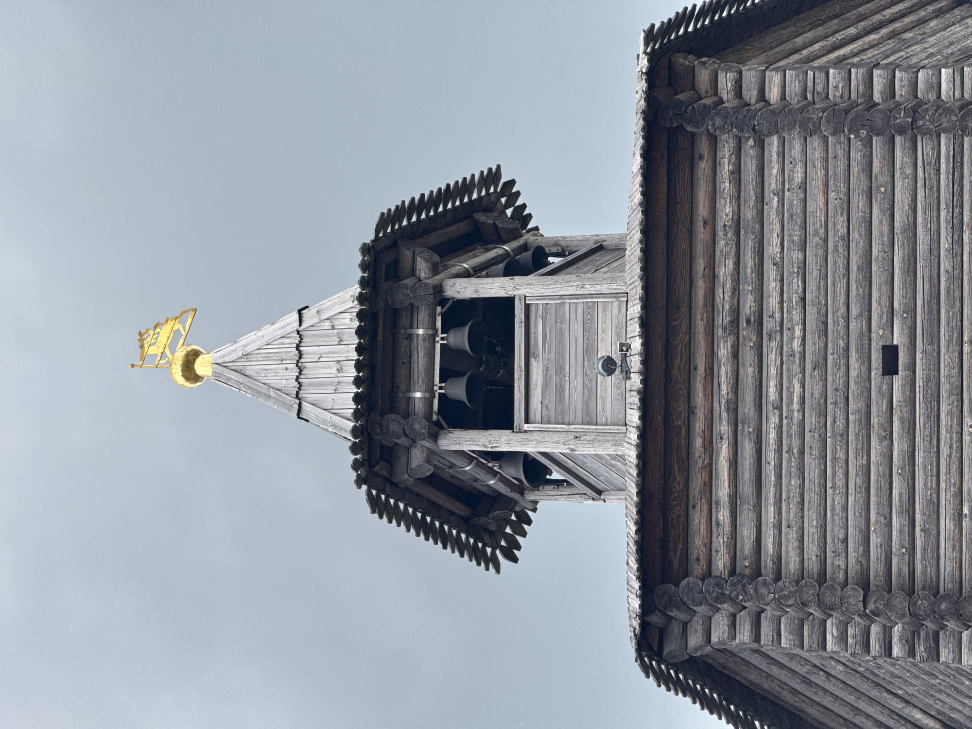 This is a low-angle, close-up shot of the top section of a wooden bell tower, likely part of a church or monastery in Nizhny Novgorod, Russia. The tower is constructed from dark, weathered logs. At the apex of the structure, a golden dome is visible, topped with a golden weather vane featuring a stylized horse-like figure. Below the dome, a tiered roof with wooden shingles leads to an open belfry where several dark, bell-shaped bells are suspended. The sky behind the tower is overcast and pale gray, suggesting a cloudy or foggy day. The image focuses on the architectural details of the wooden structure and its golden finial.