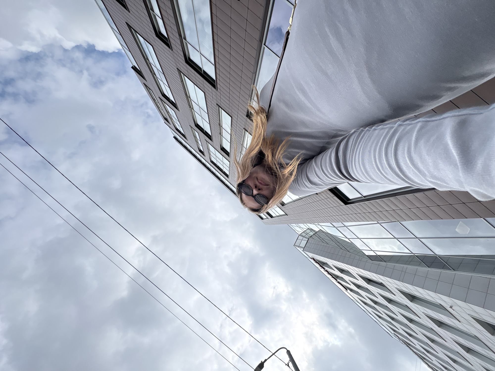 A low-angle, wide-shot photograph captures a woman from the chest up, looking upwards at the camera. She has blonde hair and is wearing black sunglasses and a light gray long-sleeved shirt. She is positioned in the foreground, with two modern buildings and a cloudy sky in the background. The building on the right has a dark brown facade with rows of windows, while the building on the left is lighter in color with a glass facade. Overhead power lines are visible against the sky, suggesting an urban environment. The overall impression is a candid shot taken on a city street.