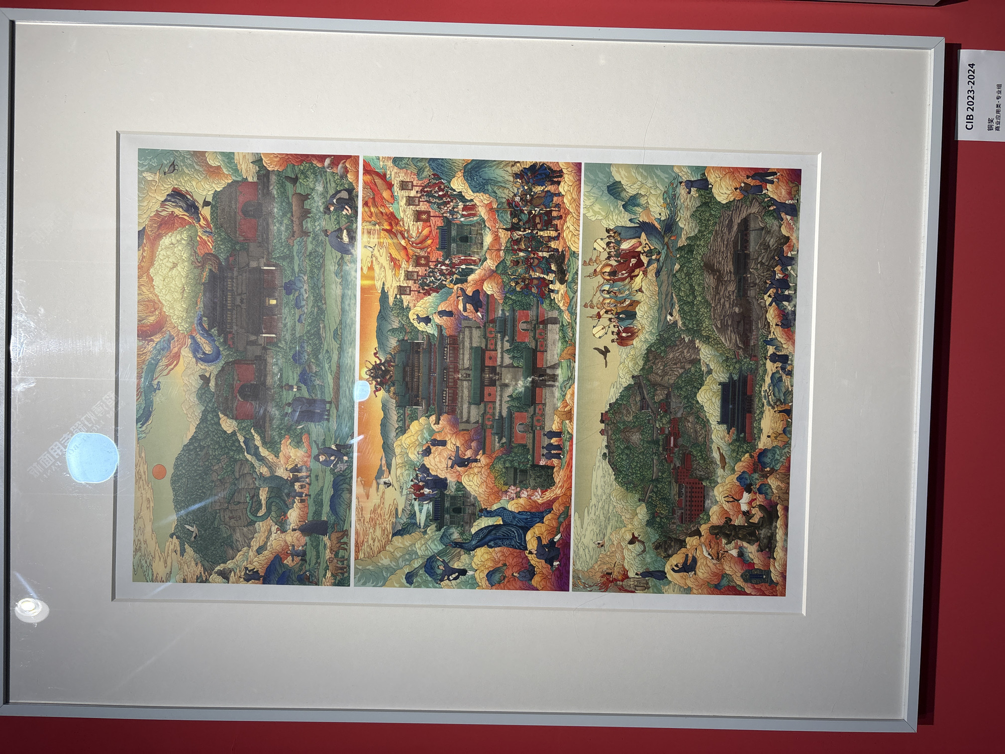 Framed artwork is displayed on a red wall. The artwork consists of three vertically oriented panels, arranged side-by-side within a single frame. The frame is white. The artwork contains an intricate, colorful illustration depicting architectural structures, landscapes, and figures rendered in a fantastical style. Buildings with traditional Asian architectural details are prominent. The figures appear to be engaged in various activities, and the landscapes suggest mountainous terrain with swirling clouds and other ethereal elements. A small sign in the upper right of the frame reads "CIB 2023-2024" in white text on a red background. Text in Chinese script is also visible on the sign. A small logo, illegible, is visible in the lower left corner of the image. The location is Nan'an District, China.