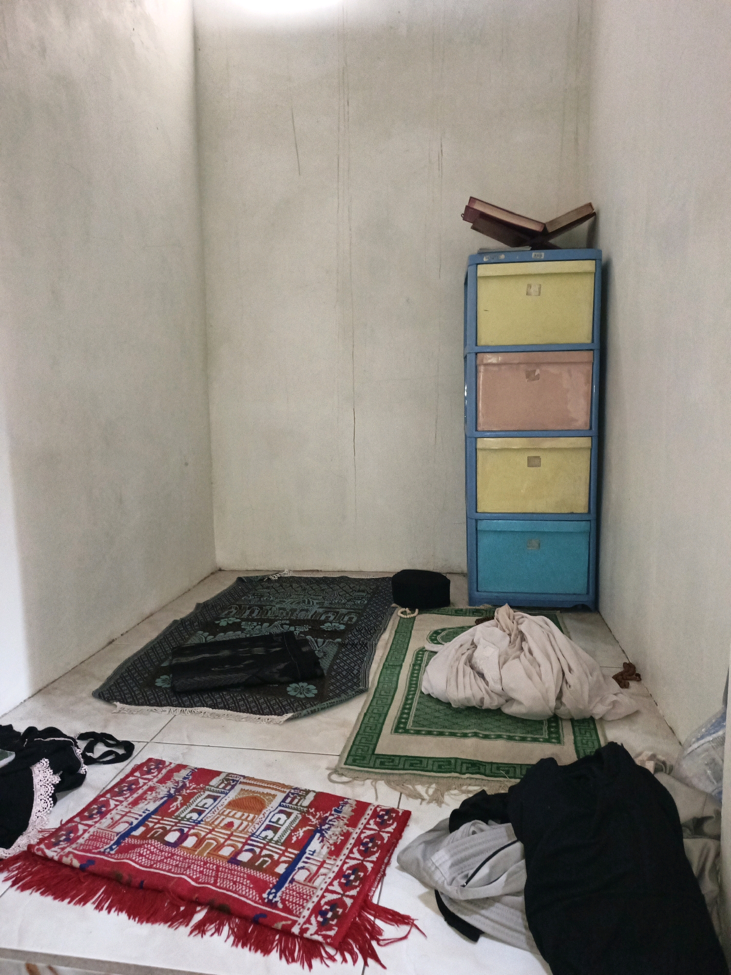 The image depicts a corner of a room, possibly a prayer room, in Ketegan, Indonesia. The walls are painted a light, textured color. In the center of the room, there are three prayer mats. The largest, a dark patterned mat, is laid out on the floor. To its right, a smaller green and white patterned mat has a pile of light-colored fabric draped over it. In the foreground, a small, vibrantly colored red prayer mat with intricate geometric patterns and red tassels lies on the floor. To the left of this red mat, a black garment with white lace trim is visible.
Against the far wall, a tall, blue plastic cabinet with four drawers of different colors (yellow, pink, yellow, and blue) is positioned. On top of the cabinet rests an open book, likely a religious text, on a book stand. A black prayer cap is placed on the floor near the cabinet. The floor is tiled with light-colored square tiles. The overall scene suggests a lived-in space used for personal devotion and rest.