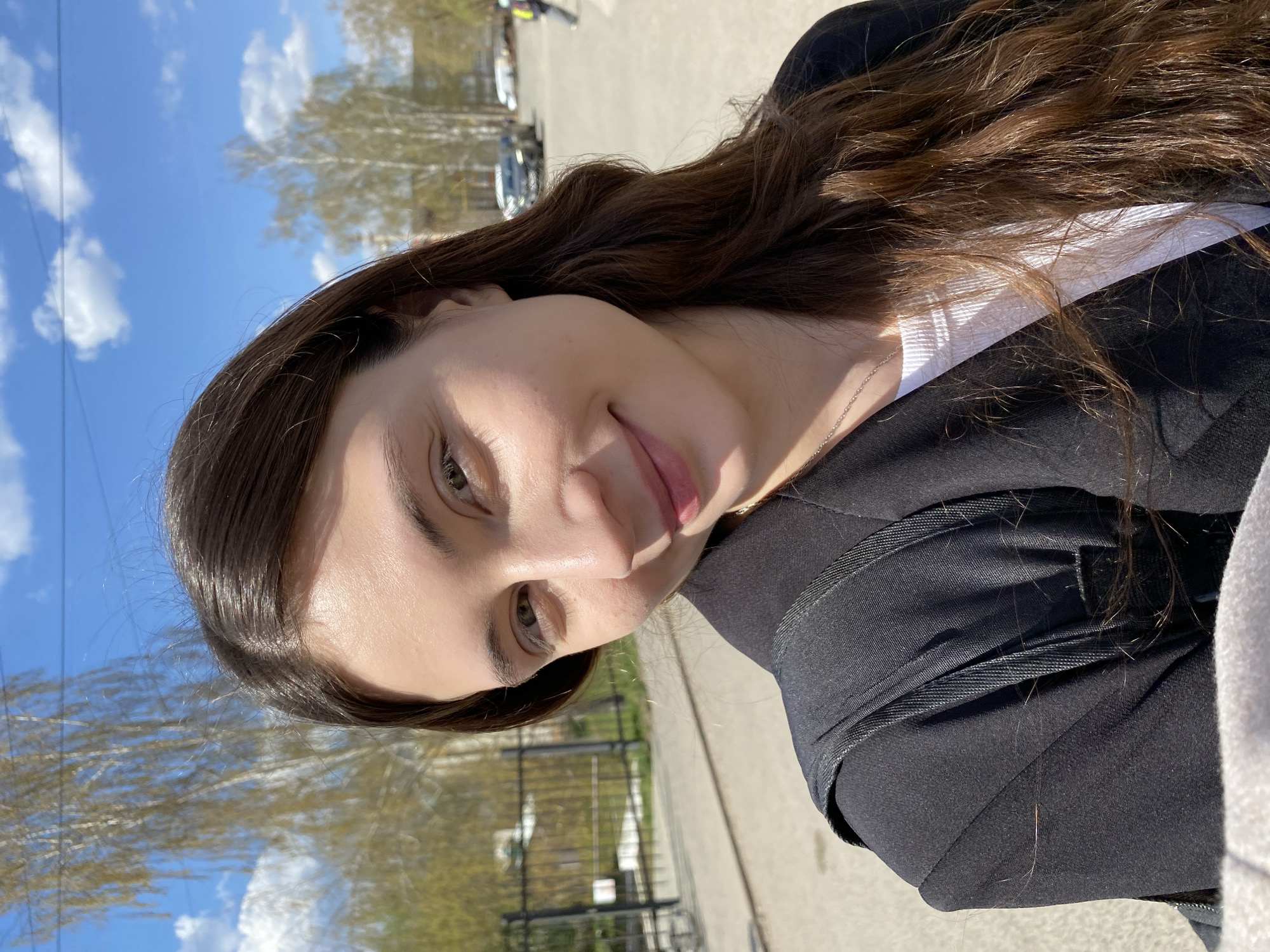 A young woman poses for a selfie outdoors in Votkinsk, Russia. The photograph is a close-up view of the woman's face and upper body, with the background showing elements of the city's environment. The woman has long, wavy brown hair. She is wearing a white top under a black jacket. The background shows a blue sky with scattered clouds, and bare trees indicating early spring. In the distance, there's a parking lot with cars and a fence, suggesting an urban park or recreational area in Votkinsk. The pavement is visible at the edge of the picture.