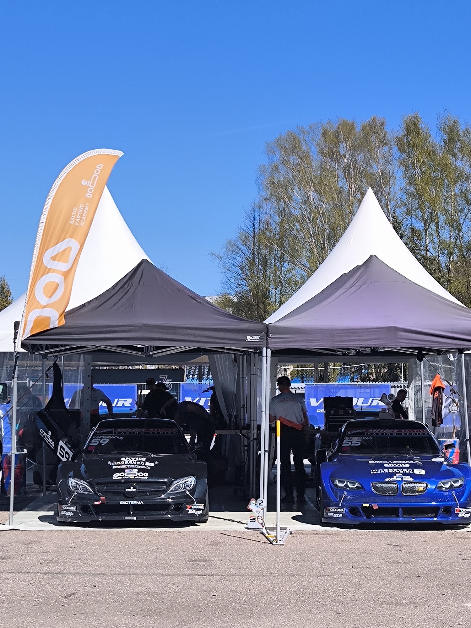 The image shows a scene likely taken at a motorsports event. Two race cars, one black and one blue, are parked under adjacent tents. The black car has the number "59" and is labelled with "Baltic Racing Series". The blue car has the number "55" and is also labelled with "Baltic Racing Series". People are working on the cars under the tents. In the background, there's a large orange and white banner that reads "Baltic Karting Academy". Further behind, there is a large white tent and trees. The ground appears to be a paved surface. The image was likely taken outdoors on a clear day. Based on the “Baltic Racing Series” and "Baltic Karting Academy" on the sign, it is most likely located in the city of Riga, Latvia.
