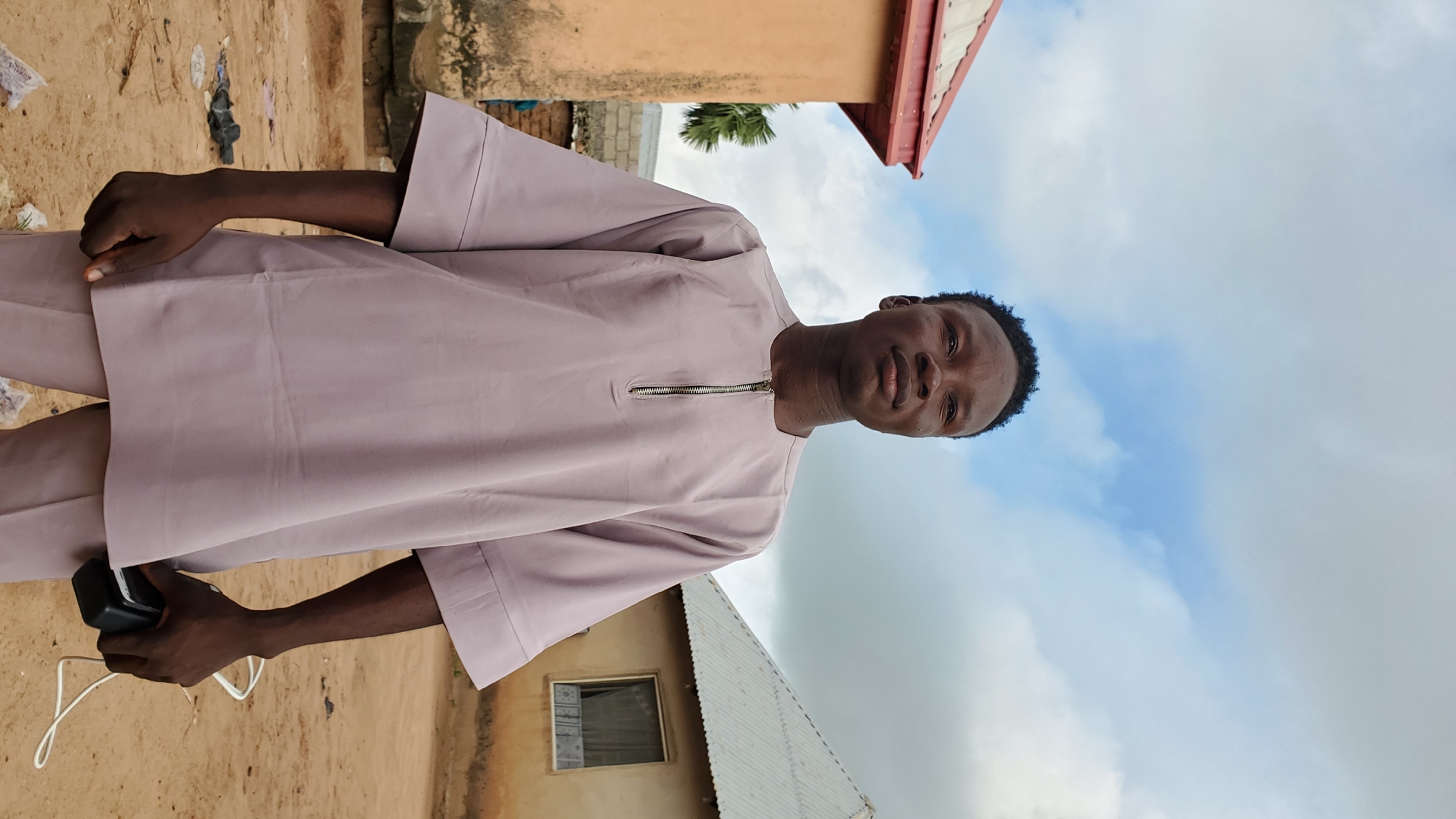 A young Black male stands outdoors, looking slightly off-camera to his right. He is wearing a light purple, loose-fitting tunic with a zipper at the neckline and wide sleeves, and matching pants. His hair is short and dark. His right hand is holding a black object, possibly a phone or a charger, with a white cord trailing from it. Behind him, to his left, is a portion of a light orange building with a red roof. To his right is a light-colored building with a corrugated metal roof and a window with curtains. The ground is a dusty, sandy surface with some scattered debris. The sky is cloudy with patches of blue visible. The overall setting appears to be a rural or semi-urban area in an outdoor environment.