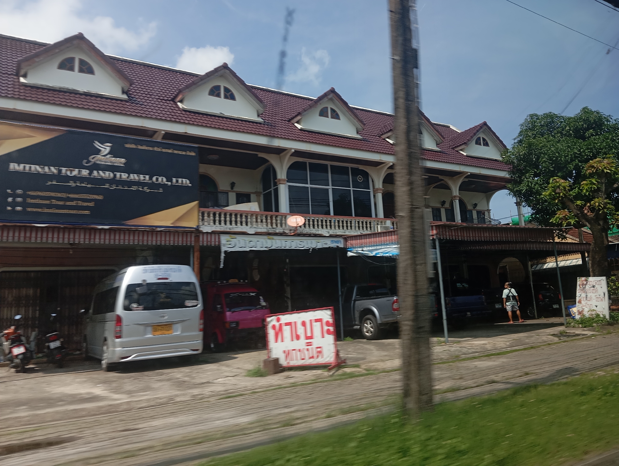 The image shows a street scene in Ko Kaeo, Thailand. In the foreground, a gray van and a pink vehicle are parked. A sign with Thai script is visible in front of the pink vehicle. The background features a building with a red tile roof and arched windows. A person wearing a light-colored shirt and shorts stands near the building. Several trees and power lines are also present in the scene. The sky is blue with some clouds.
