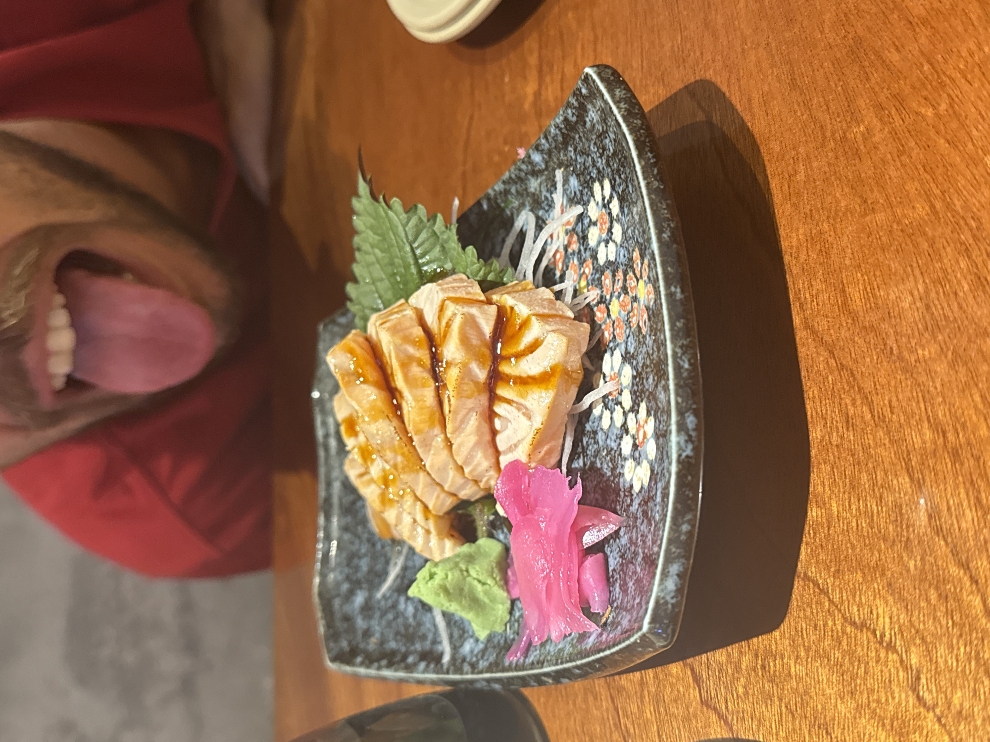 A plate of seared salmon sashimi, served with shredded daikon radish, a green shiso leaf, wasabi, and pickled ginger, sits on a wooden table. A person wearing a red shirt is visible to the left of the plate, with their mouth open and tongue extended. The plate is dark-colored with a floral design. The location is likely a restaurant in Đà Nẵng, Vietnam.