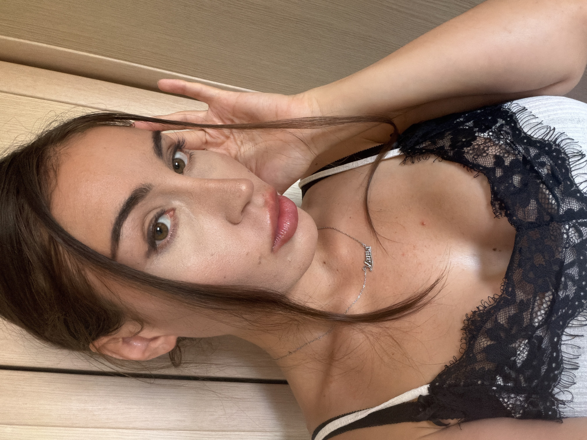 A close-up, overhead shot captures a young woman with dark hair, fair skin, and brown eyes. She is wearing a black and white lace camisole and a silver necklace with a script pendant. Her right hand is raised to her face, touching her hair, as she gazes at the camera. The background consists of a light-colored, textured wall. The scene appears to be indoors, possibly in a residential setting in Tersefanou, Cyprus.