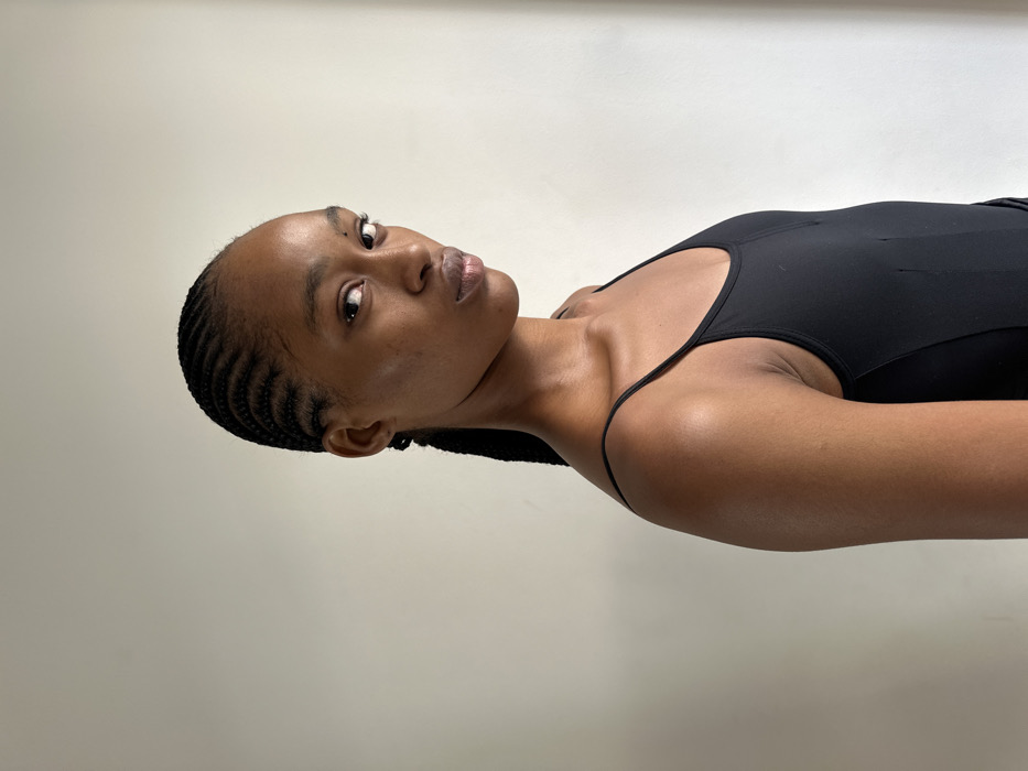 A portrait-style photograph captures a single female subject from the mid-chest upward against a plain, light-colored wall. The subject is an adult with medium-dark complexion and black hair styled in tight cornrow braids that extend into a single braid or ponytail behind her head. She wears a black, thin-strapped garment, possibly a tank top or bodysuit. Her body is angled slightly to the viewer's right, while her head is turned to face the camera directly, establishing eye contact. Her expression is neutral. Minor skin texture is visible on her face, and a small dark mark is present on her left cheekbone. The lighting is uniform and diffused. No other individuals, objects, or environmental details are present to indicate the location within Paris, France, or any other geographical area.