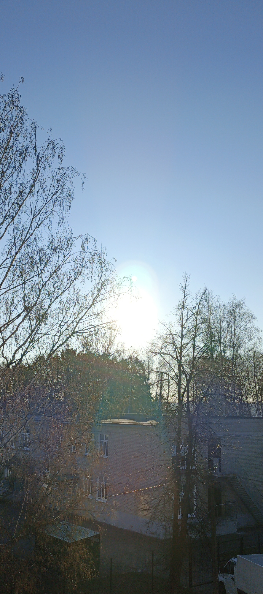 The image displays an outdoor scene in Chaikovsky, Russia, taken from an elevated viewpoint. The upper half of the frame is dominated by a clear, bright blue sky. The sun is visible slightly to the right of the center, creating a strong lens flare and illuminating the scene.
Numerous bare deciduous trees, possibly birches given the visible catkins, feature prominently in the foreground and mid-ground, their intricate branch patterns silhouetted against the sky. A dense line of trees, including some evergreens, forms a dark band across the horizon, situated behind the more immediate bare trees.
Below the tree line, a multi-story building with light-colored brick or stucco walls and numerous rectangular windows is visible. It appears to be a residential or institutional structure with a flat roof section. To the right, another part of a building shows an external metal staircase leading upwards. A small, covered structure is partially visible on the ground level to the left. A fence line runs horizontally across the lower part of the image, and a white truck is partially visible in the bottom right corner.
No people or specific activities are depicted. The strong, low-angle sunlight suggests either an early morning or late afternoon setting. The bare trees indicate that the season is likely late autumn, winter, or early spring. The weather is clear and sunny. There is no visible text within the image. The scene conveys a calm, natural-urban environment on a bright day.