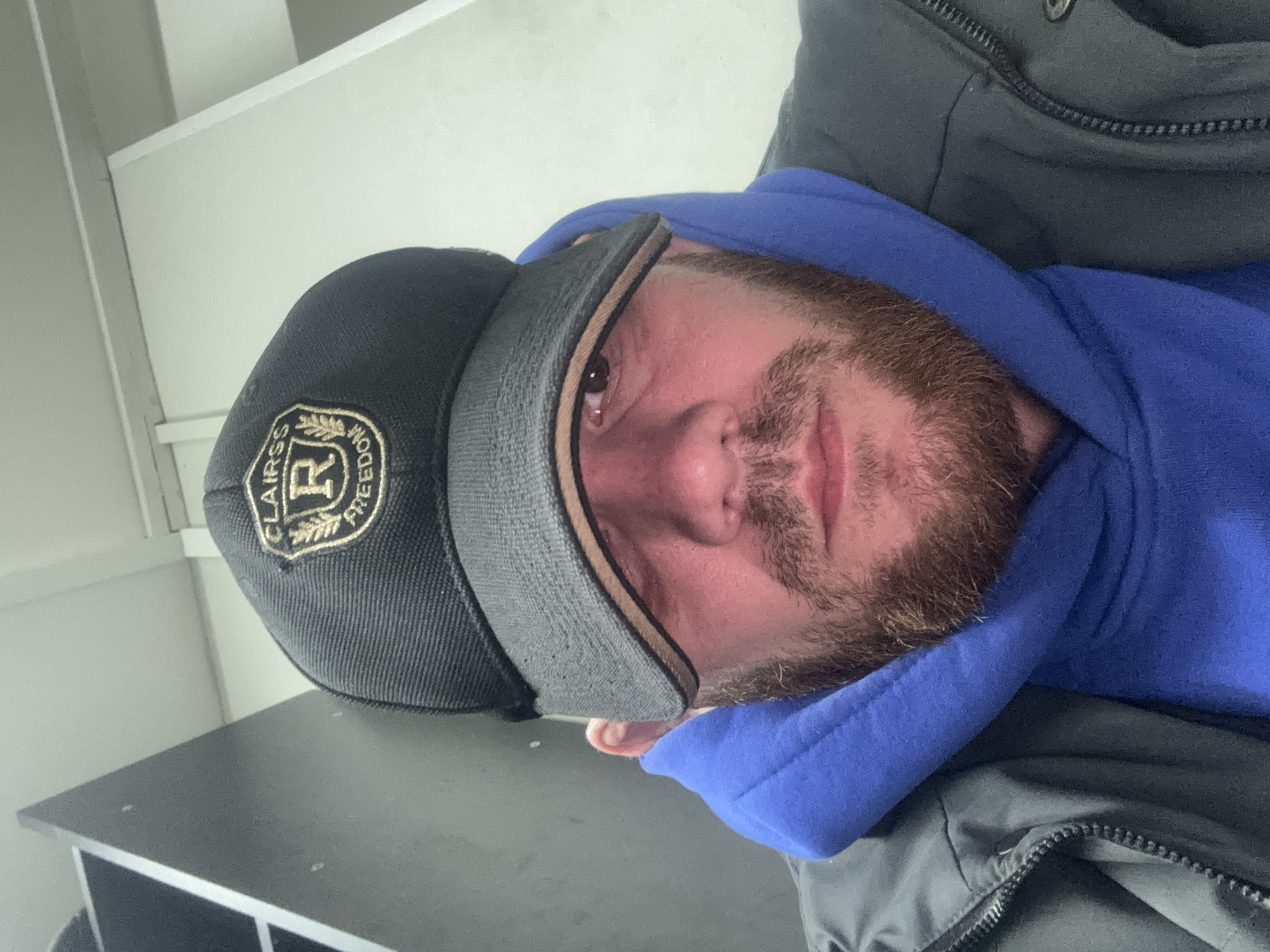 A man with a red beard and wearing a black cap with a patch and a blue hoodie is in a gray jacket, presumably indoors. The location is unknown, and there is no information to suggest that the image was taken in Perm, Russia. The man is likely taking a selfie. A table or shelf is visible in the background.
