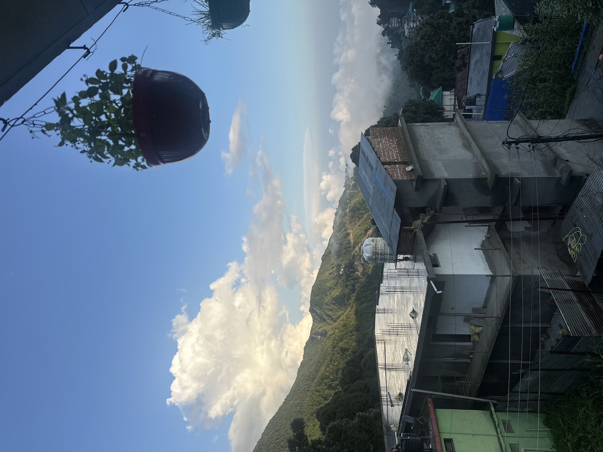 The image depicts a mountainous landscape with buildings and a cloudy sky. In the foreground, there is a partially constructed building with exposed rebar and concrete on its roof. Adjacent to it, another building is visible, painted green. Several other buildings with varied roof colors are scattered across the hillside. Overhead, a hanging potted plant with green foliage is suspended, adding a touch of nature to the scene. The background features lush green mountains shrouded in mist, with a vast expanse of sky dotted with white clouds. The overall impression is a serene, rural setting.