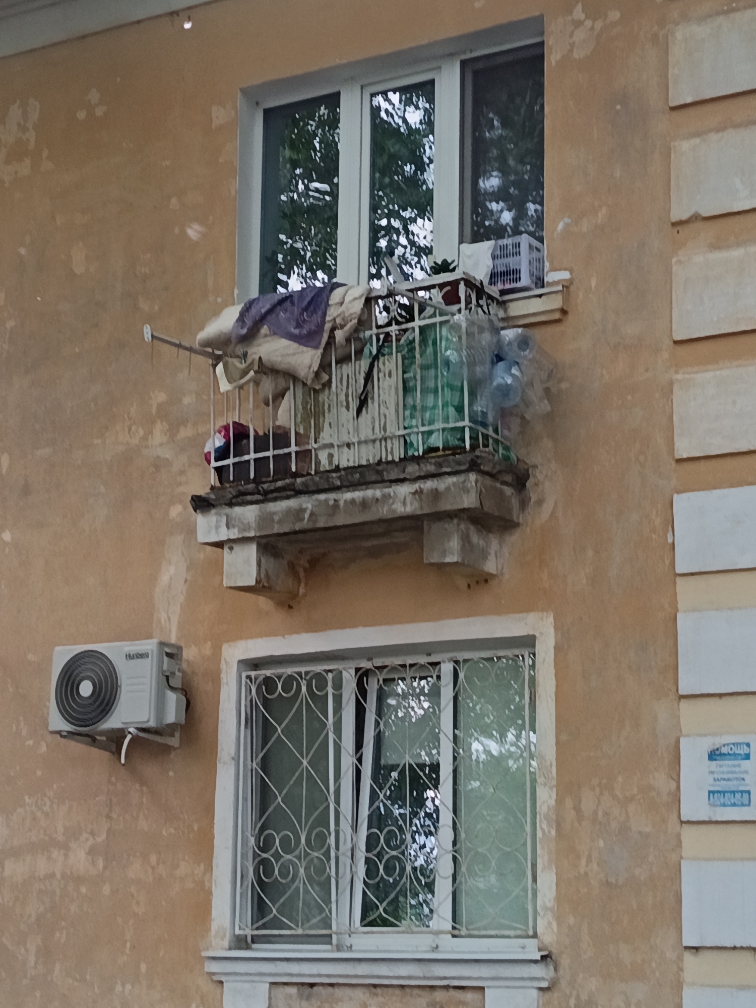 This is a medium-distance shot of a tan, weathered building facade, likely an apartment complex, in Komsomolsk-on-Amur, Russia. The building features two windows and a small, enclosed balcony. The upper window is a large, white-framed casement window with multiple panes, revealing greenery from trees outside. Above the balcony, a clothesline extends from a rod on the wall to the balcony railing, with some garments hung on it, including a dark purple piece of fabric and lighter colored cloths. The balcony itself is enclosed by a white metal railing and contains several objects: a white plastic crate, what appears to be a plant in a pot, a green and white striped bag, and several clear plastic bottles. The balcony is supported by a concrete structure that shows signs of age and deterioration.
Below the balcony, to the left, is a light gray air conditioning unit mounted on the wall. To the right of the AC unit is a second, smaller window, also white-framed, with decorative white metal grillwork featuring swirling patterns. This window also reveals greenery through its panes.
The wall of the building has patches of peeling paint, indicating it is an older structure. The overall lighting suggests it is daytime, possibly overcast due to the soft light. On the right side of the building, a small blue and white sign with Russian text is partially visible. The visible text includes "ПОМОЩЬ" (POMOSHCH, meaning "HELP") and a phone number beginning with "8 (800)...". The scene conveys a sense of lived-in residential architecture, with the balcony items suggesting personal use and the overall condition of the building pointing to its age and location in a possibly industrial or older urban area.