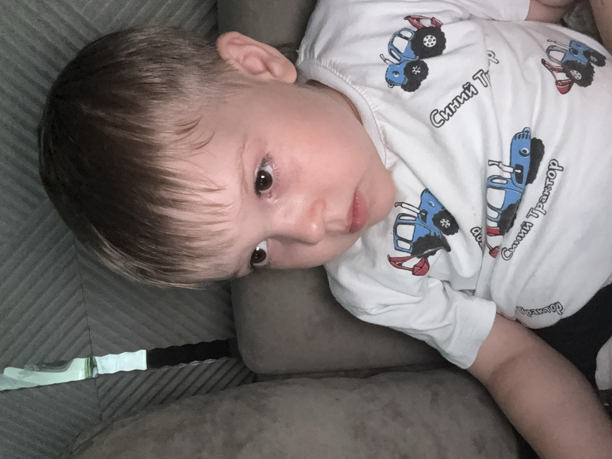 A young boy with light brown hair is looking to his left, with his mouth slightly open, while sitting on a sofa. He is wearing a white t-shirt with blue tractor graphics. The t-shirt has text in Russian that reads "Синий Трактор" (Blue Tractor). The background consists of a gray sofa and a striped, dark gray fabric. The image was taken in the city of Oktyabrskoye, Russia.