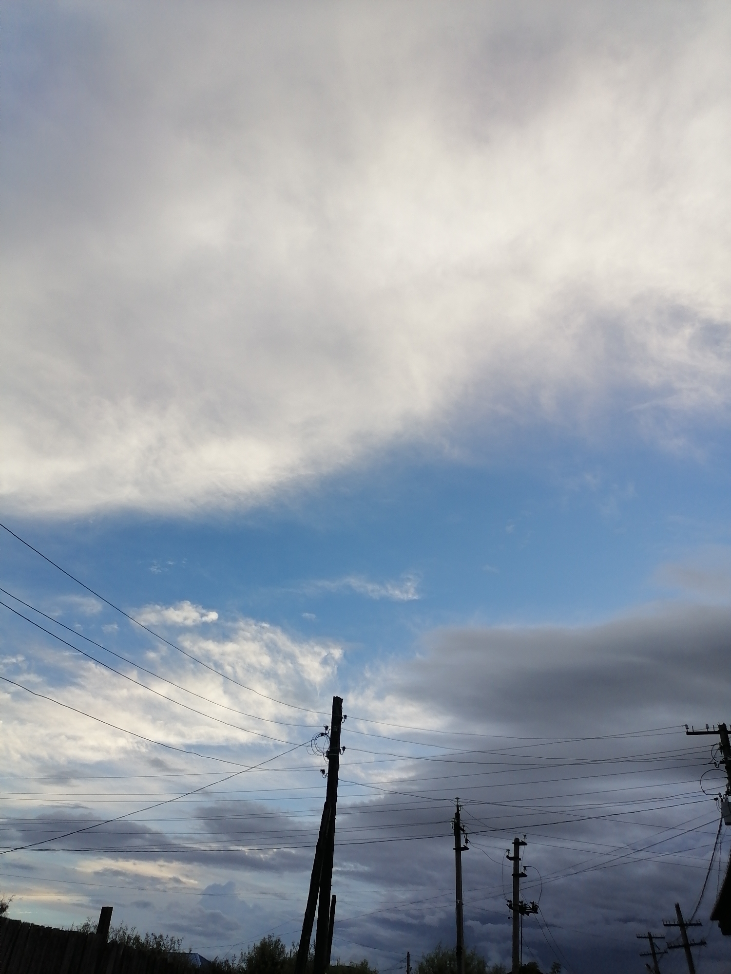 The image depicts a cloudy sky with power lines and utility poles in the foreground. The sky transitions from bright white clouds at the top to a darker, more ominous gray cloud formation near the horizon. The blue sky is visible in the middle band. Several power lines crisscross the frame, supported by wooden utility poles, some of which are silhouetted against the sky. The ground level shows a dark fence on the left and sparse vegetation. The scene appears to be outdoors, likely in a semi-urban or rural setting. There are no people or identifiable buildings visible, other than a hint of a roof structure on the far right.