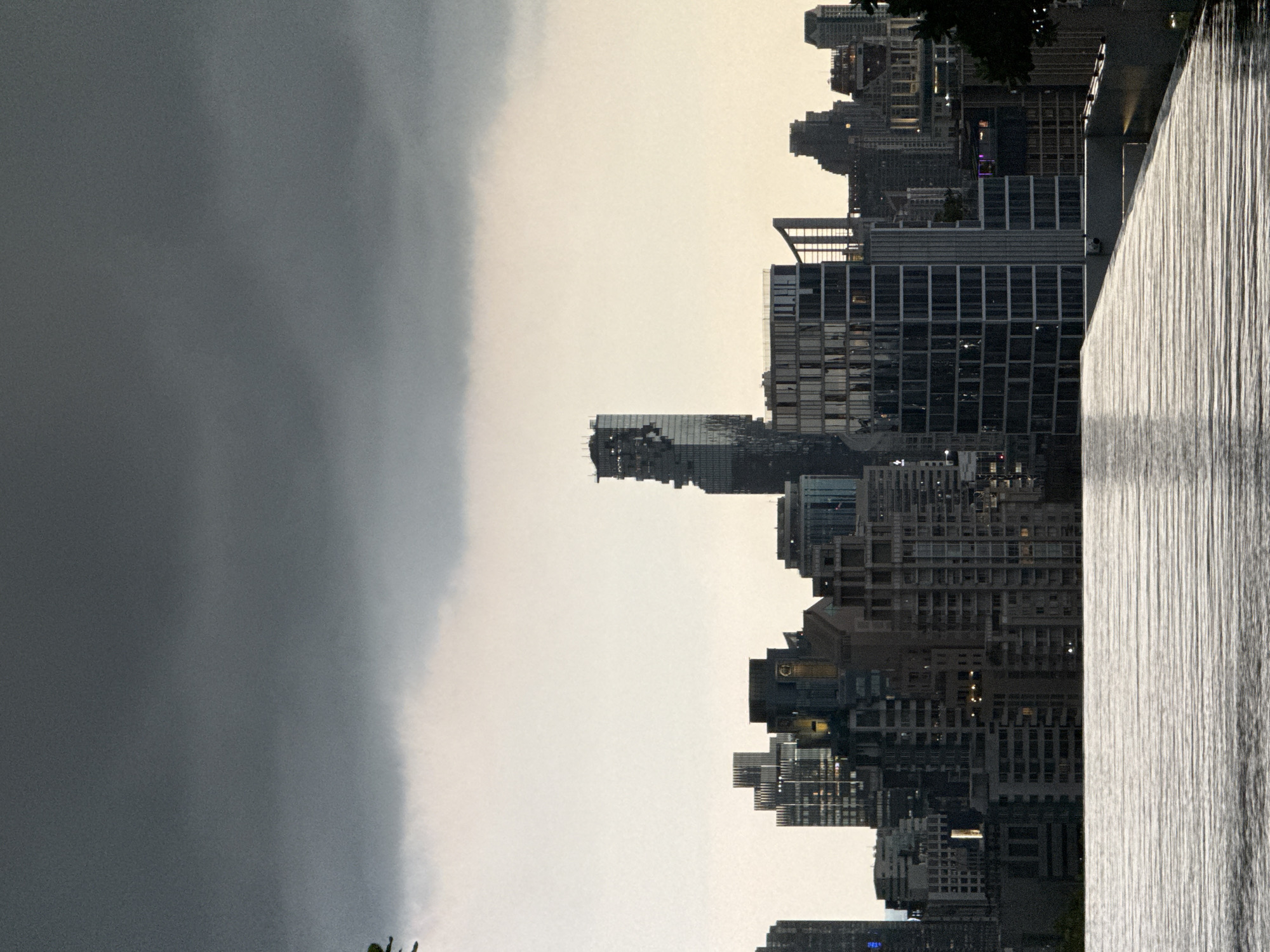 The image shows a skyline of Bangkok, Thailand, under a cloudy sky. High-rise buildings dominate the cityscape, with a large body of water or a water feature in the foreground, potentially part of a public park or development. The city appears to be experiencing stormy weather on one side, indicated by the dark clouds. The lighting suggests it could be dusk or dawn.