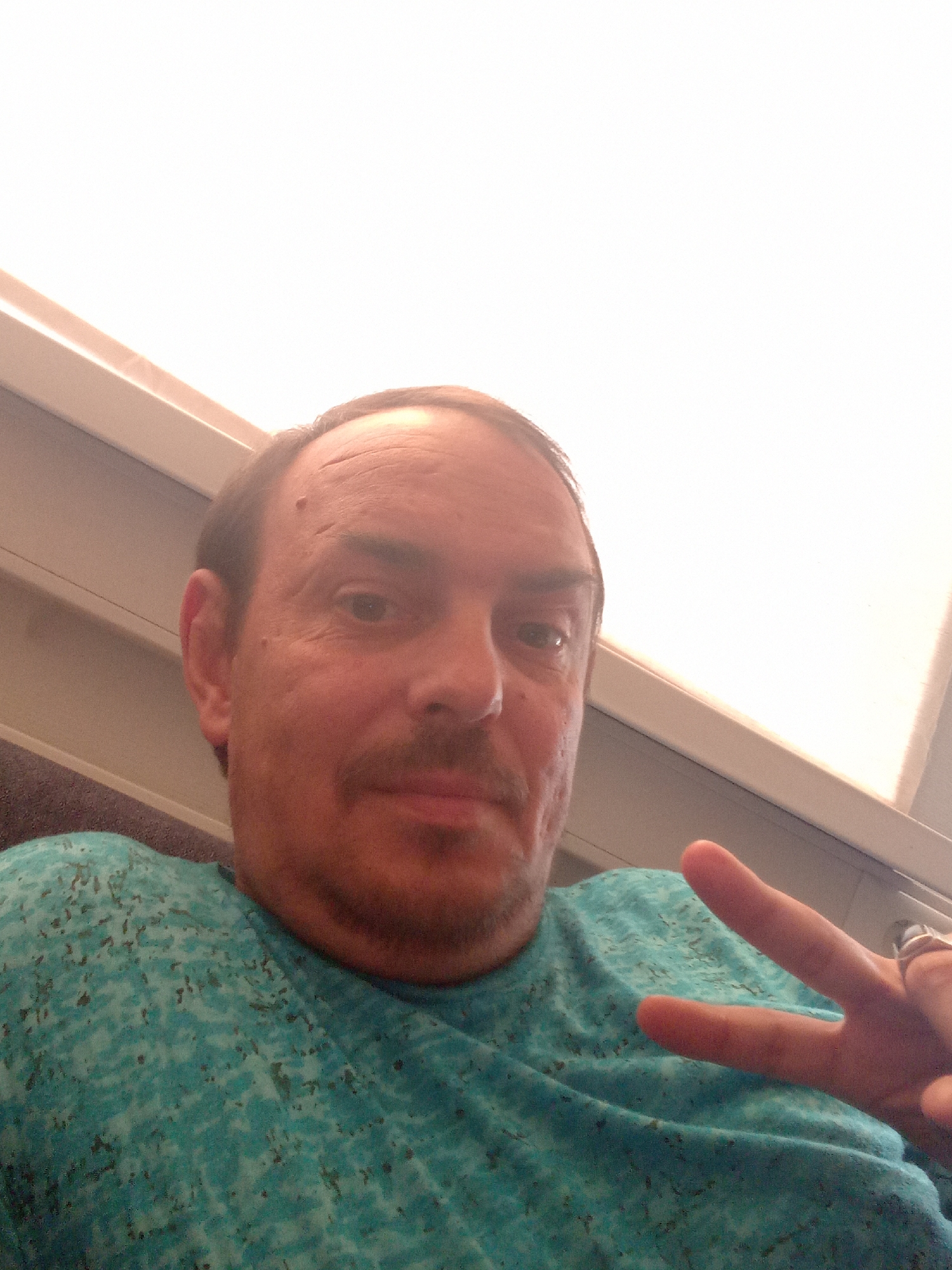 A close-up, waist-up, selfie-style photograph captures a light-skinned man with thinning brown hair, a mustache, and a mole on his forehead. He is wearing a teal t-shirt with a speckled pattern. The man is positioned in the foreground, with his head slightly tilted. His right hand is raised, forming a peace sign, and he is wearing a ring on his index finger. The background is out of focus, but appears to be an interior space with a white ceiling or wall and some beige trim. The lighting is bright, suggesting it might be daytime. There is no indication of the city of Selli, Estonia, in the image itself.
