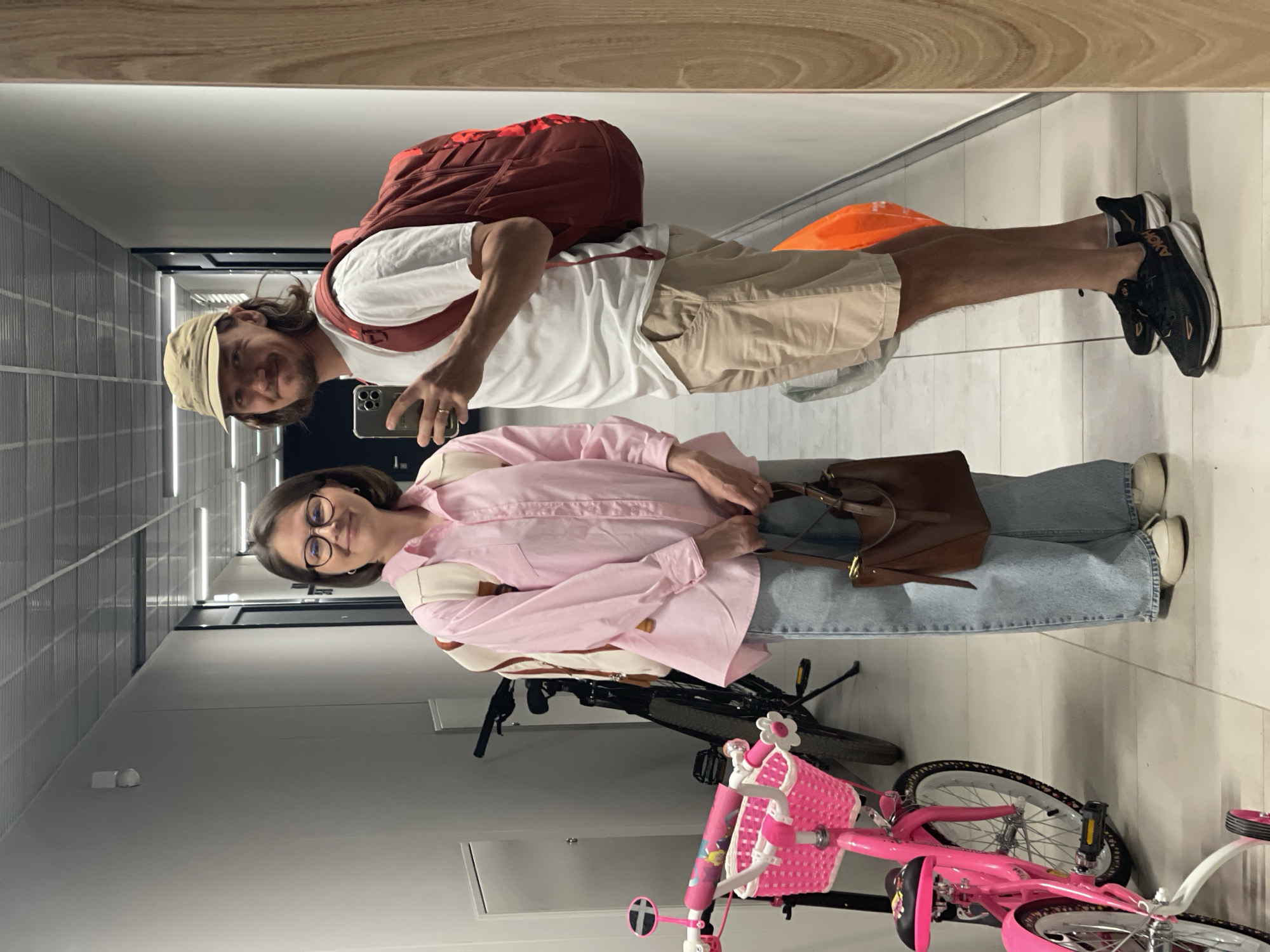 The image is a selfie taken in a hallway. A man and a woman stand side-by-side, apparently in front of a mirror, as evidenced by the mirrored image quality. The man wears a white T-shirt, khaki shorts, a tan baseball cap, and black running shoes. He has a red backpack. He holds a smartphone in his right hand, presumably to take the picture. The woman wears a pink button-down shirt, light-wash jeans, and glasses. She carries a brown handbag.
Two bicycles are visible in the foreground. One is a pink child's bicycle with a basket. The other is a black adult bicycle. The hallway has gray walls and a tiled ceiling. The lighting appears to be artificial. There are no features that would definitively indicate that this image was taken in Izhevsk, Russia.