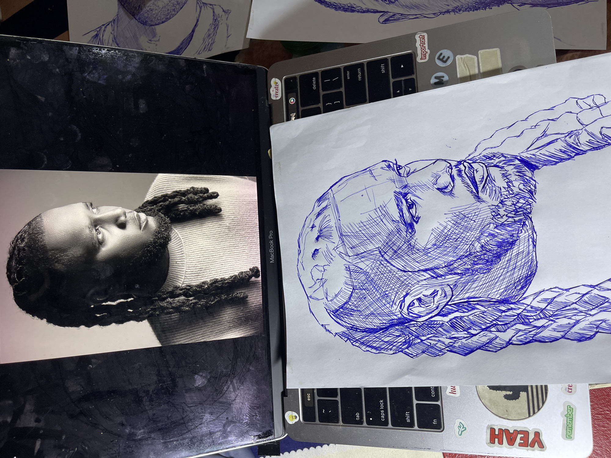 The image displays a partially completed blue ink drawing of a man's head and shoulders, laid across a laptop keyboard. The drawing is on a white sheet of paper and appears to be a sketch, with various cross-hatching techniques used to create shading and texture. The man in the drawing has braided hair, a beard, and is looking to the right. The laptop is a MacBook Pro, visible by the branding on the bottom bezel. The keyboard keys are discernible, including 'esc', 'tab', 'caps lock', 'shift', 'fn', 'control', 'delete', and 'enter'. Several stickers are affixed to the laptop casing, including one with a lightbulb icon and another that says "creates". A portion of a photograph or screen displaying a black and white image of the same man from a similar profile view is visible on the laptop screen, suggesting the drawing is a portrait study. The scene is lit from above, casting shadows. The environment is a flat surface, likely a desk, with the edge of another drawing visible on the right side of the image. The image does not contain any elements that can be used to determine its location in Jos, Nigeria.