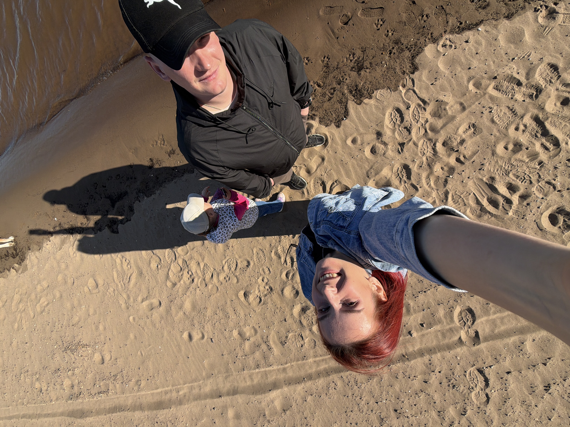 A high-angle, full-body shot depicts three individuals standing on a sandy area adjacent to a body of water, potentially the Kama River or one of its tributaries, in the city of Chaikovsky, Russia.
On the left side of the frame stands a man wearing a black Puma baseball cap, a black jacket, and dark trousers. He is looking up at the camera.
To the right of the man is a small child wearing a white hat and a patterned shirt. The child also looks at the camera.
Closest to the camera, a woman with red hair is holding her arm outwards, presumably taking the selfie. She is wearing a denim jacket.
The sandy ground is covered in various footprints, suggesting recent activity. The upper left corner of the image shows the edge of the body of water. The scene is well-lit, suggesting it is daytime.