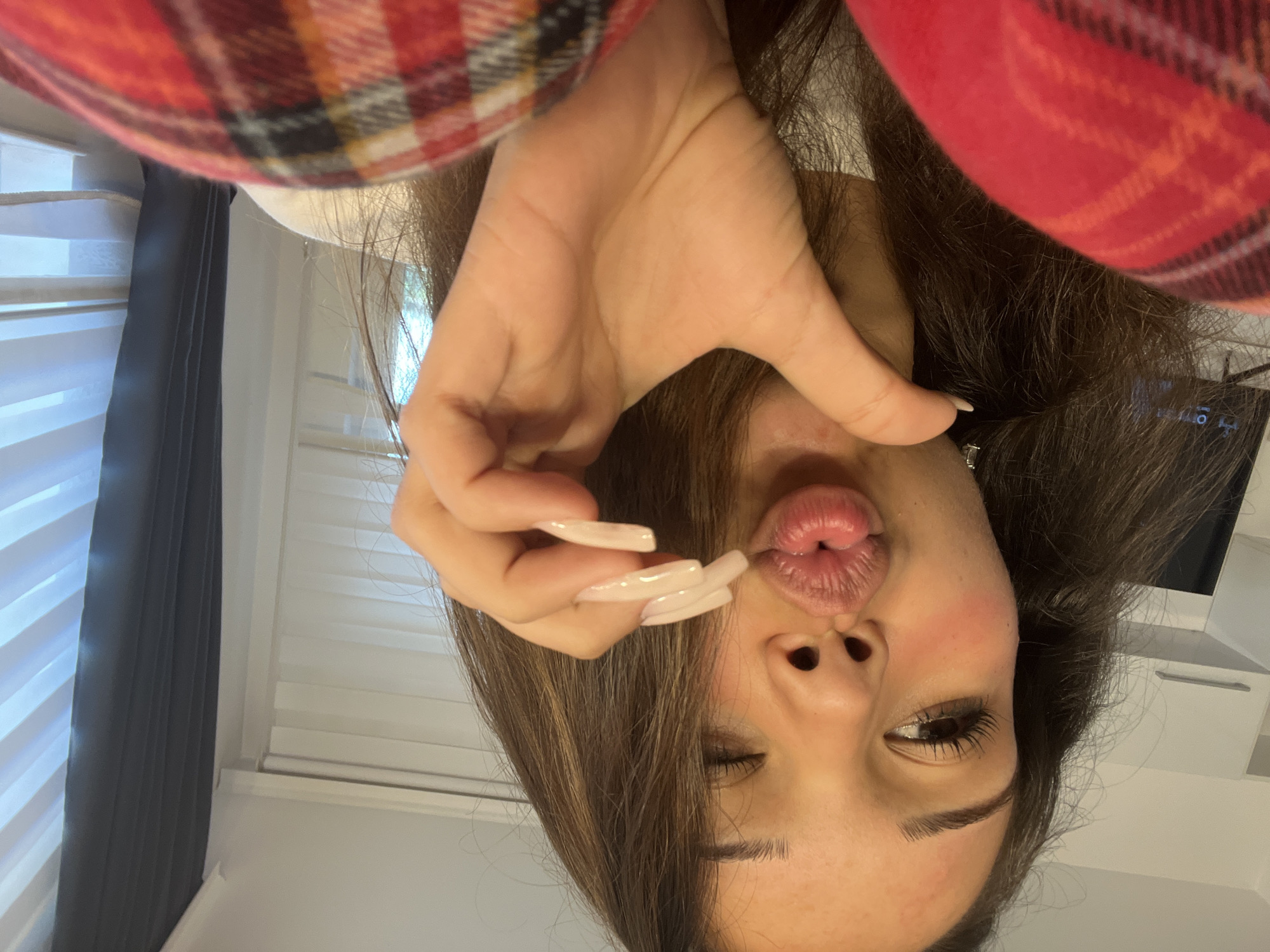 The image shows a close-up, head-and-shoulders view of a young woman with long, brown hair, posing for a selfie. She is making a kissing face, with her lips puckered, and is holding up her hand with long, light-colored fingernails in front of her face. She is wearing a red and black plaid garment on her left shoulder. The background appears to be an indoor setting with blinds and dark curtains on a window. There is no indication of the city of Zlatograd, Bulgaria, in the image or its context.
