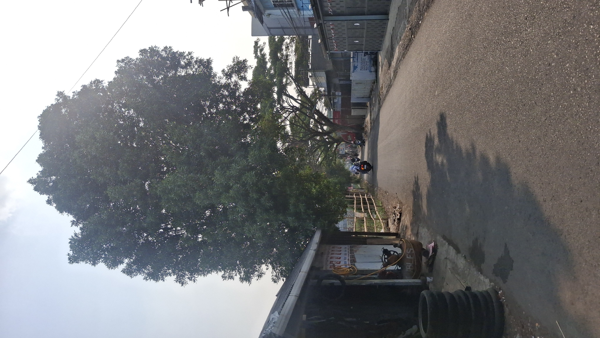 A tree-lined, paved road in Tasikmalaya, Indonesia, under a bright sky. The road is bordered on the left by a low, corrugated metal-roofed structure with a wooden fence in front of it. Piles of stacked tires are visible next to the structure. Several motorcycles are traveling down the road, some carrying passengers. On the right side of the road, there are more buildings with dark gray decorative panels and a blue-sided building further down. Large trees with dense green foliage dominate the scene, casting shadows on the road. The overall impression is of a somewhat rural or suburban street on a clear day.