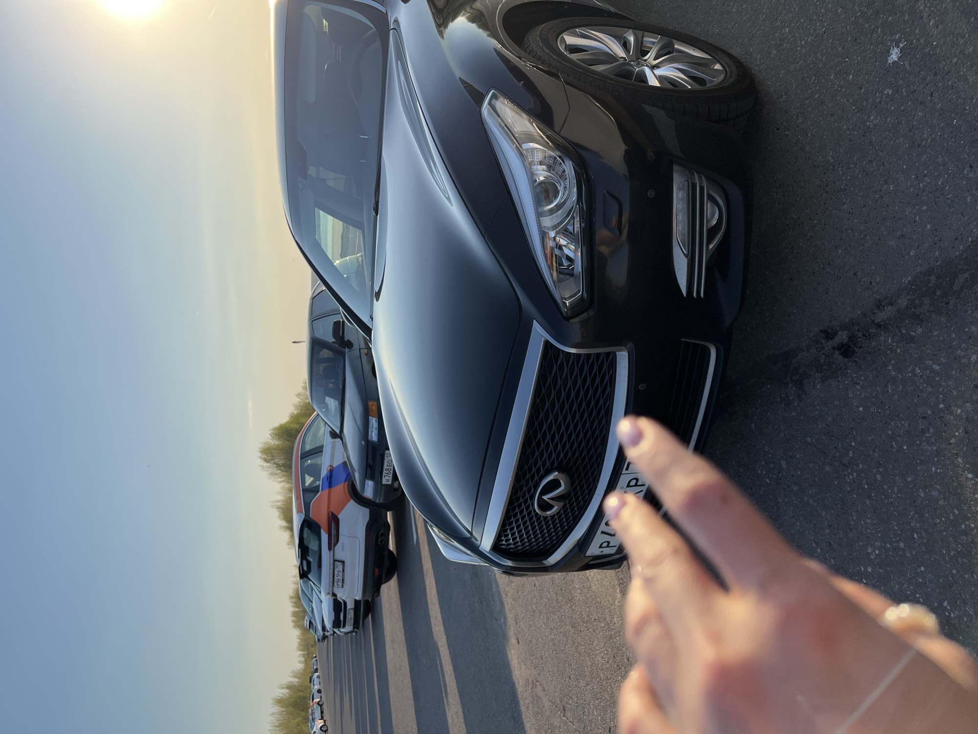The image shows the front of a black Infiniti car parked on an asphalt surface, likely a parking lot or a road. Several other cars are parked behind it in a row. The lighting suggests either early morning or late afternoon. The scene appears to be outside, possibly near a road or open area. A hand is partially visible in the foreground. The location is indicated as Sviyazhsk, Russia.