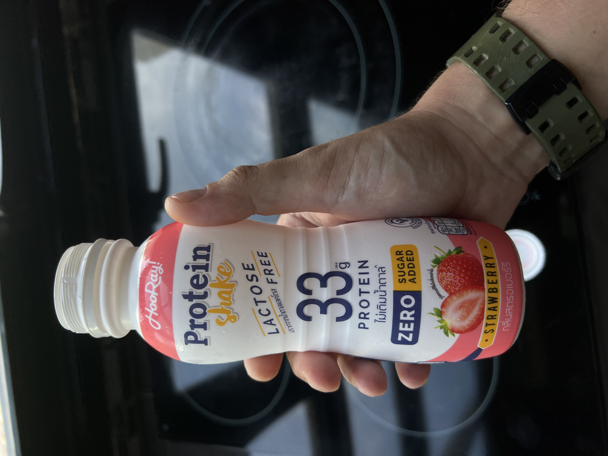A hand wearing a green watch strap holds a bottle of "HooRay! Protein Shake" strawberry flavored drink. The bottle is white with pink and blue labeling. Prominently displayed are the words "Protein Shake," "LACTOSE FREE," "33g PROTEIN," and "ZERO SUGAR ADDED." Below this, there is an image of two strawberries and the word "STRAWBERRY." The background appears to be a dark surface, possibly a stovetop, with faint circular markings. The image is taken from a slightly low angle, focusing on the bottle and the hand holding it.