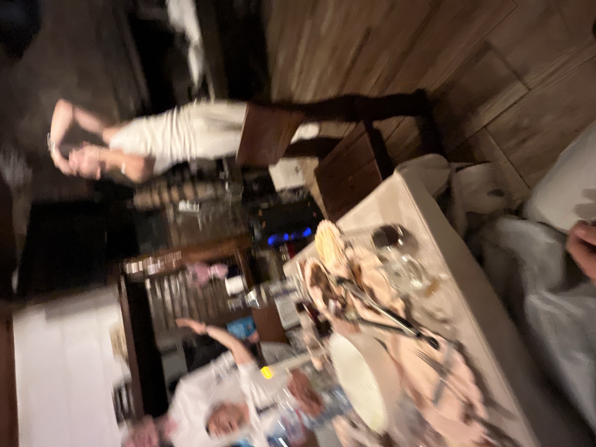 Indoors at a restaurant, presumably in Presoka, Bulgaria, the scene shows two people dining at a table. One person, wearing a white shirt, is partially visible at the lower left edge of the frame, with one arm raised. Another individual is seated at the head of the table, leaning back in their chair. The table is set with plates, cutlery, glasses, and napkins. The surrounding environment appears to include dark wooden floors and a cabinet, along with other furniture and interior details typical of a dining space. The image is blurry, possibly indicating motion during the photo's capture.
