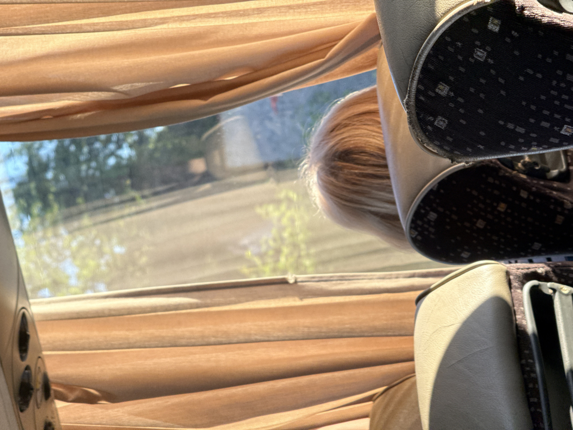 The image shows the interior of a bus with peach-colored curtains pulled back, revealing sunlight streaming through the windows. In the foreground, two bus seats are visible. The seat on the left is light beige and appears to be an armrest. The seat on the right has a dark fabric with a subtle pattern. Behind the seats, a person's blonde hair is visible, suggesting they are sitting on the bus and looking out the window. The background shows a blurred outdoor scene with trees and greenery, indicative of a natural environment. This scene is consistent with being inside a bus traveling through or parked near Kiosevtsi, Bulgaria, a village known for its natural surroundings.