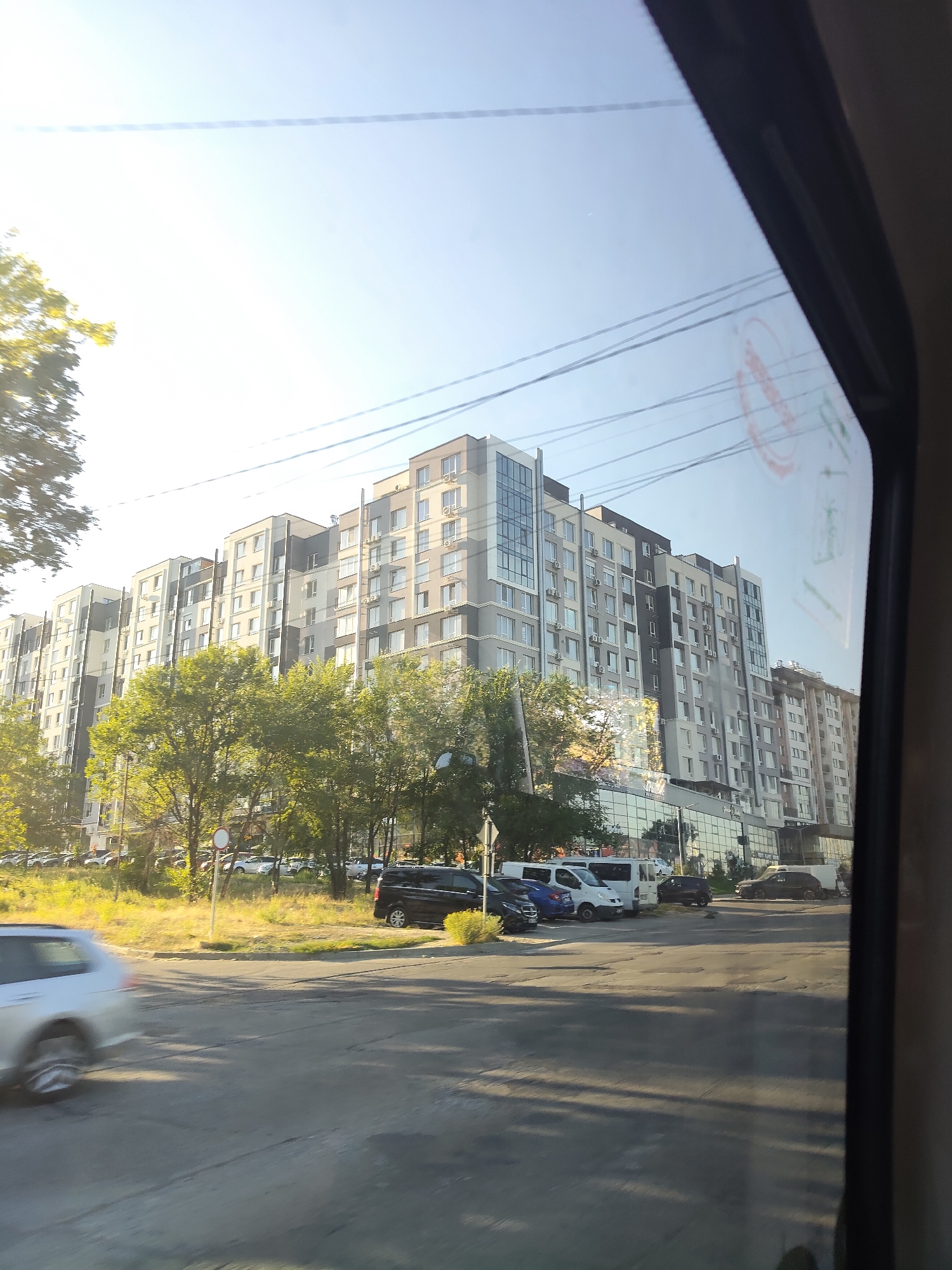 The image shows a street view in Chișinău, Moldova, with a large, modern apartment building as the dominant structure. The building features a grey and white facade with numerous windows and balconies, and appears to be several stories high. In front of the building, a line of trees with lush green foliage is visible. Cars are parked along the street and in a lot in front of the building, including a silver station wagon in the foreground, a black SUV, and several vans. The street itself is paved and appears to be an intersection or a busy road, with cars driving. Overhead, power lines are strung across the sky, which is clear and bright, suggesting a sunny day. The reflection of what appears to be a red circular sticker or sign is visible on the right side of the image, likely on the window through which the photo was taken. The overall scene depicts a typical urban residential area with elements of modern architecture and daily life.