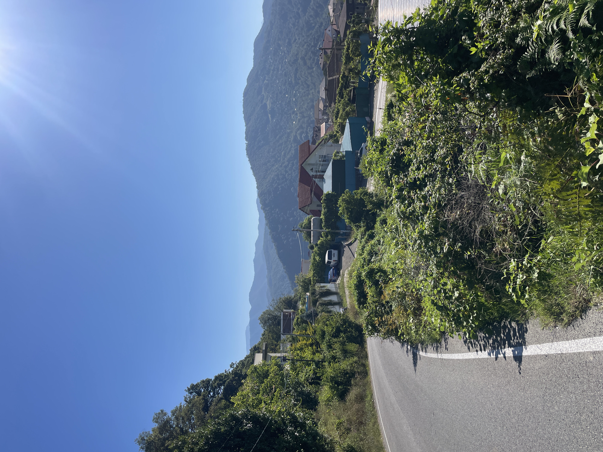 The image depicts a sun-drenched, mountainous landscape in Baranovka, Russia. A winding asphalt road, marked with a white line, curves through lush green vegetation on the left side of the frame. On the right side of the road, several houses with red and blue roofs are nestled among the hills, surrounded by trees and dense foliage. In the distance, layers of mountains recede into a clear blue sky. The sun is shining brightly, casting shadows on the road and illuminating the scene. There are no people visible in the image. The overall impression is one of a serene, rural setting with evidence of residential development.