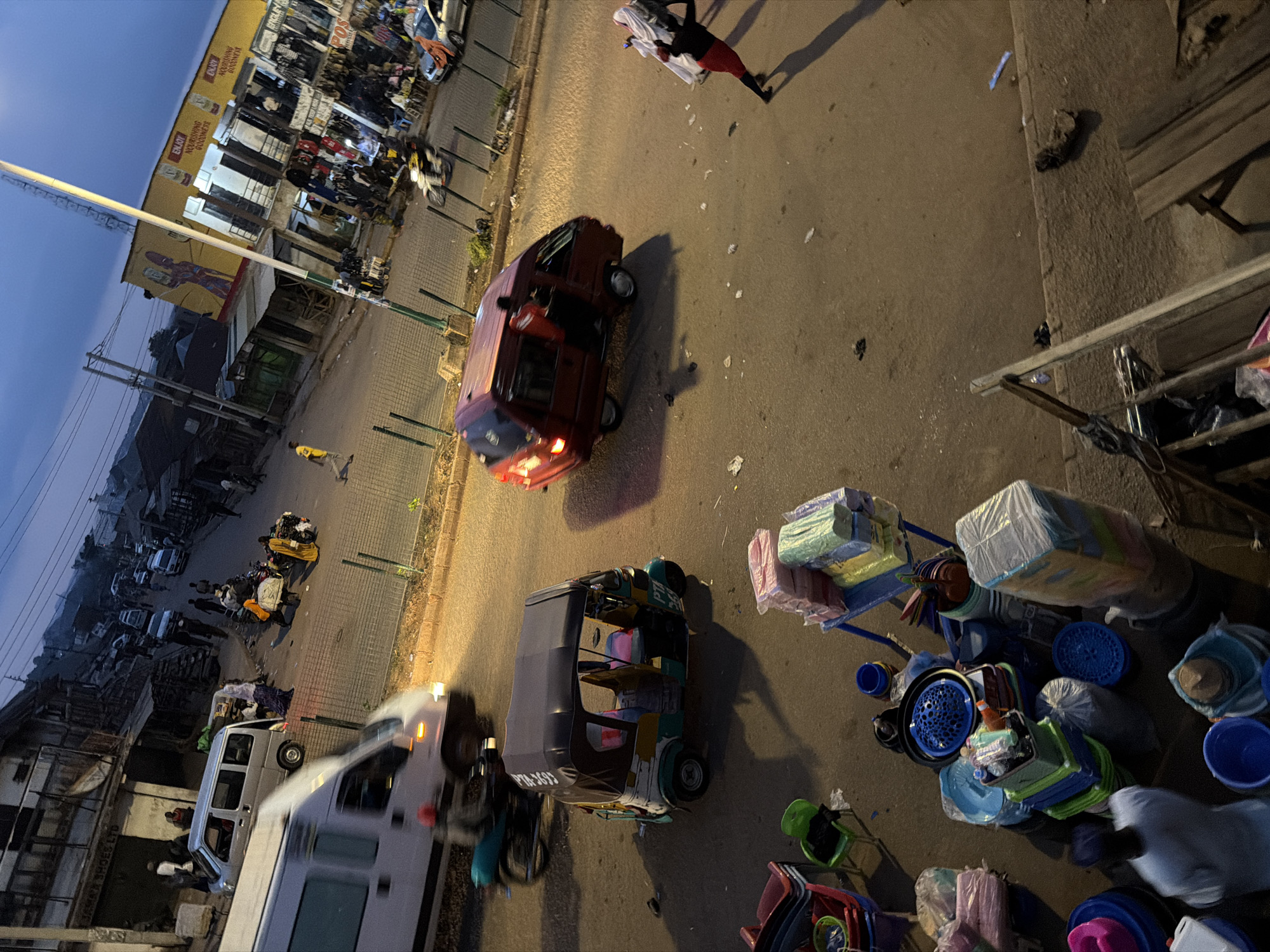 The image depicts a busy street scene in Bukuru, Nigeria, during twilight. Several vehicles are present, including a white van in motion on the left, a red van with its door open in the center, and a three-wheeled auto-rickshaw parked on the right. Pedestrians are scattered throughout the scene, with some walking and others gathered near shops. A person wearing a red shirt and black pants is walking away from the red van, carrying a white bundle on their head. Numerous goods, including stacked plastic containers, buckets, and packaged items, are displayed on the sidewalk and on carts, suggesting a market or commercial area. The buildings lining the street appear to be shops, some with awnings and signage. Streetlights are illuminated, casting a warm glow on the pavement. The overall atmosphere is one of daily life and commerce in a Nigerian urban setting.