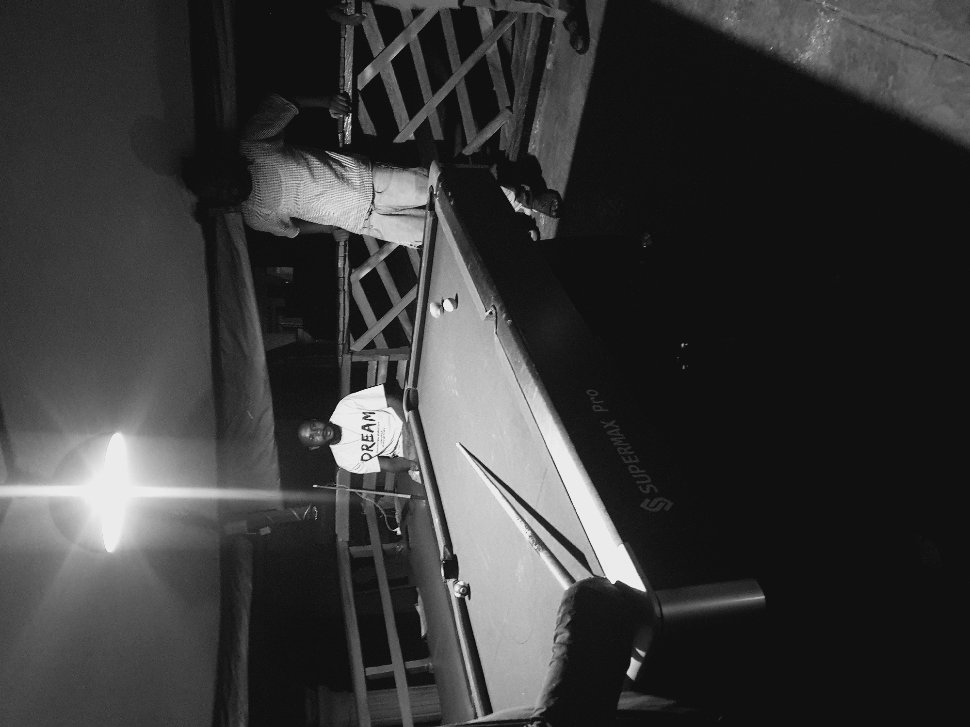 The black and white image shows a dimly lit outdoor scene at night, possibly on a balcony or deck, with a pool table as the central focus. Three adult males are visible. One man, wearing a t-shirt with the word "DREAM" printed on it, sits on a stool at the far end of the pool table, looking towards the camera. Another man, wearing a checkered shirt and shorts, stands leaning on a wooden railing behind the pool table, facing away from the camera. A third man's legs and torso are visible in the foreground on the left, appearing to be about to play pool. Two white pool balls are resting on the table near the man in the checkered shirt. A cue stick lies across the table in the foreground. A bright overhead light fixture illuminates the scene from above. The background consists of a wooden railing and a dark, possibly natural, environment. The word "SUPERMAX Pro" is visible on the side of the pool table. The scene suggests a casual gathering for a game of pool in Lokoja, Nigeria.