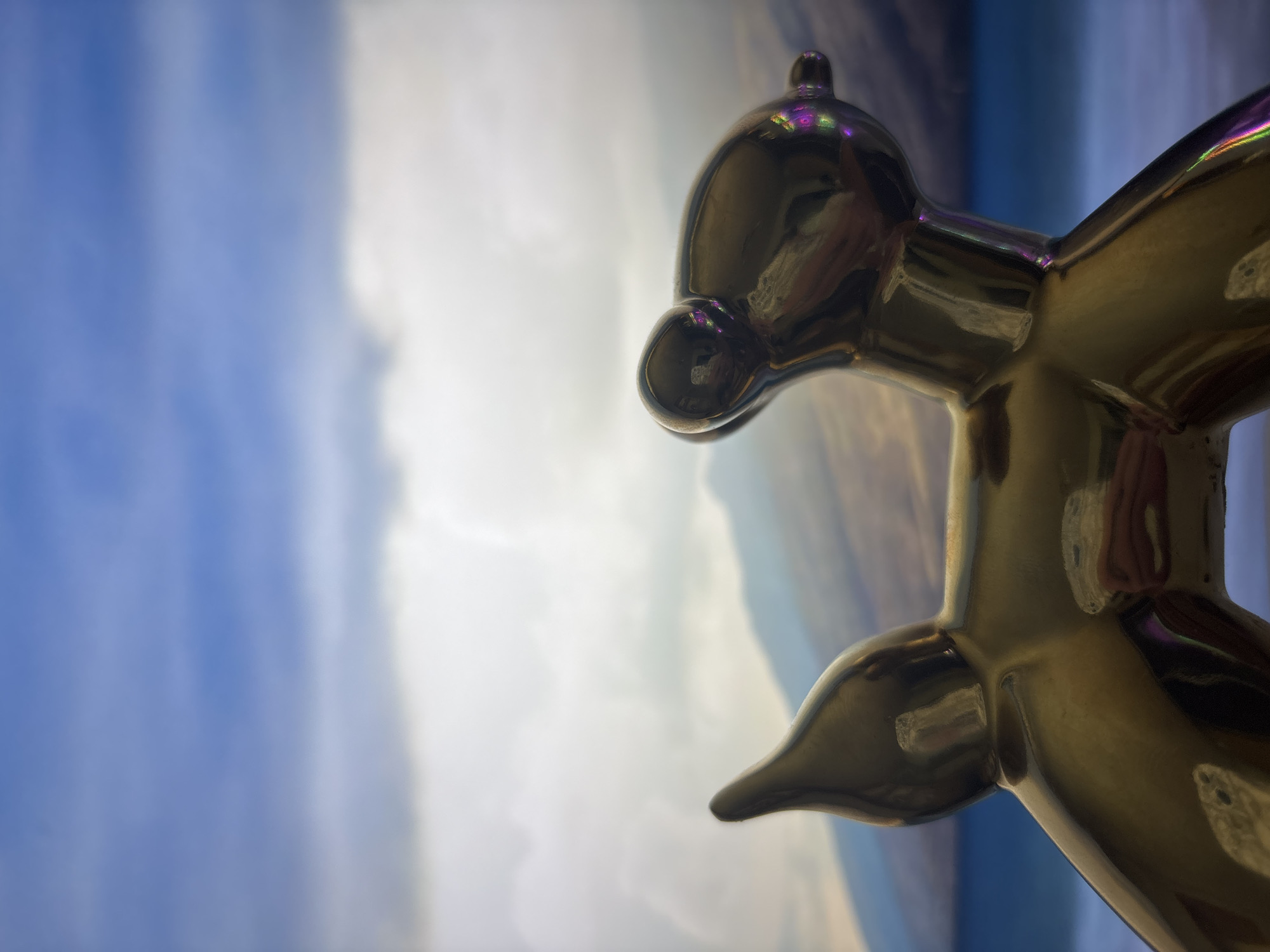 This media file is a close-up, low-angle shot of a shiny, metallic sculpture resembling a balloon dog. The sculpture is golden in color and has a highly reflective surface that mirrors the background. The background itself appears to be a blurred, abstract depiction of a sky with variations of blue and white, suggesting clouds or a painted backdrop. The lighting is bright, causing highlights and reflections on the sculpture. The sculpture's form is detailed, showing the characteristic curves and rounded shapes of a balloon animal. The focus is sharp on the sculpture, particularly its head and torso, while the background is intentionally out of focus, creating a sense of depth and emphasizing the subject. There are no people visible in the frame, nor is there any discernible text. The overall impression is one of contemporary art, with the reflective surface of the sculpture interacting dynamically with the soft, atmospheric background.