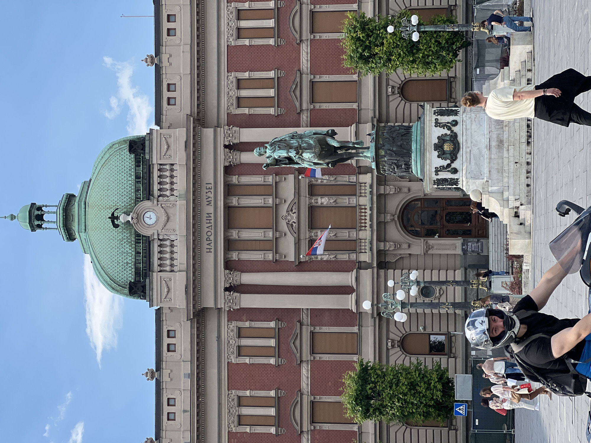 The image shows the National Museum of Serbia in Belgrade. The building's facade is visible, featuring brickwork, columns, windows, and a dome on the left. A statue of Prince Mihailo stands in front of the building. People are walking nearby, including a person wearing a helmet. A Serbian flag is flying on the building. There are trees and lamp posts in front of the museum. The sky is partly cloudy.