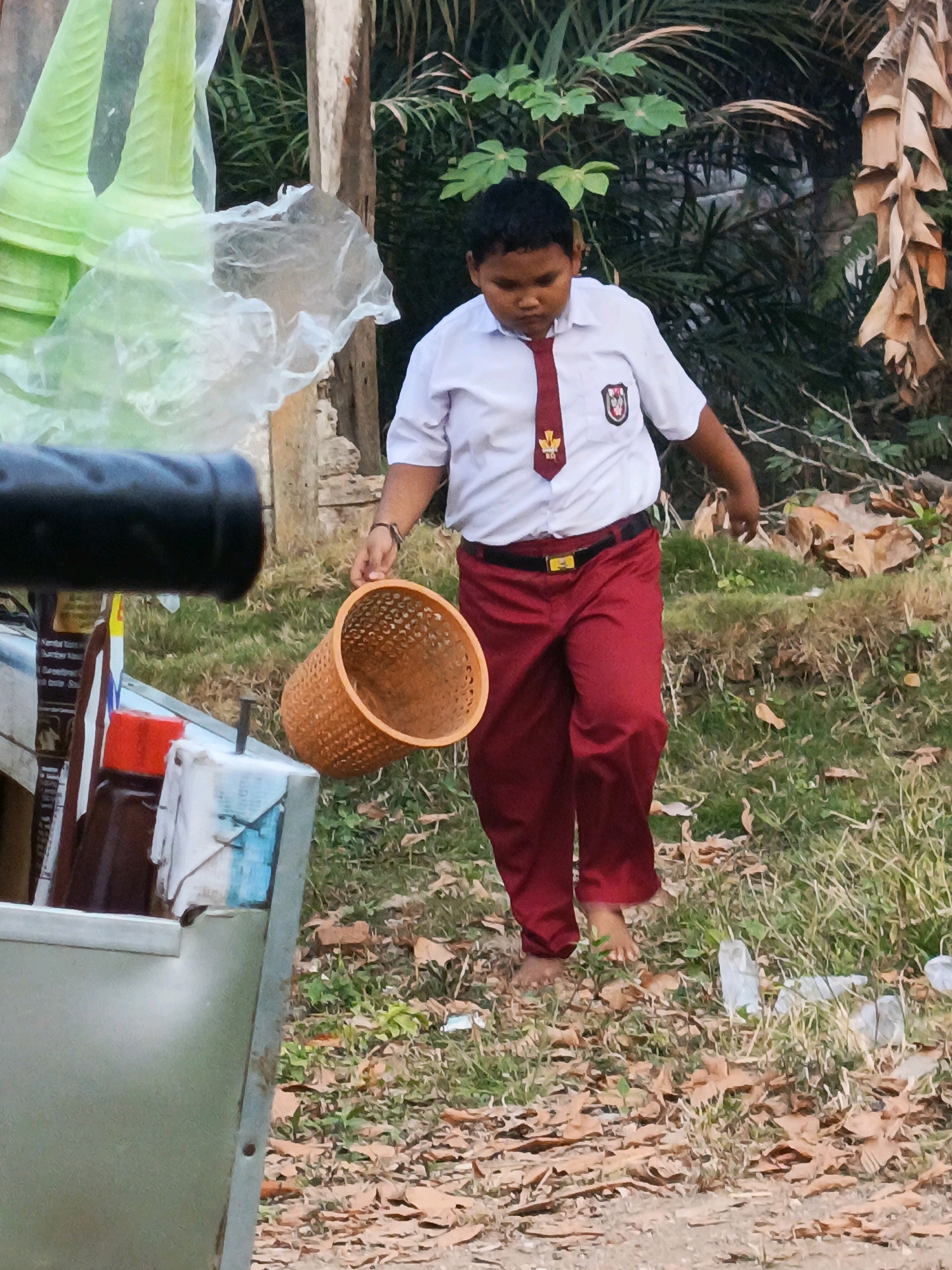 A young boy wearing a white shirt, red tie, and red pants, with a school emblem on his shirt, is walking barefoot on a grassy area. He is holding an empty woven basket in his right hand. In the background, there are trees, dried leaves, and a structure that appears to be made of stone and wood. To the left of the boy, there is a metallic structure with various items on it, including bottles and plastic bags. The scene is set outdoors, likely in a village or rural area, given the vegetation and the boy's attire. The location is identified as Megang Sakti II, Indonesia.
