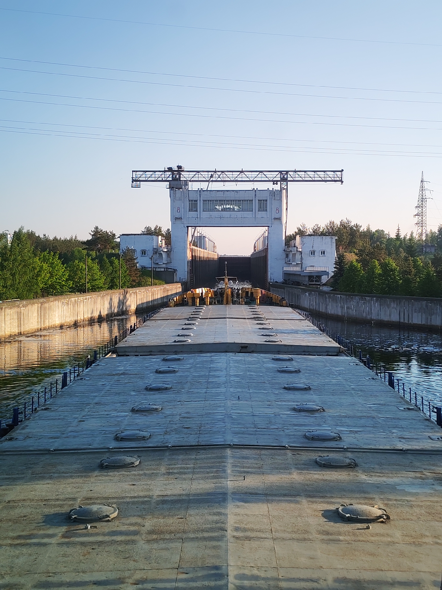 The image depicts a barge moving through a canal towards a large structure, likely a lock system. The canal is bordered by concrete walls, and dense vegetation is visible on either side. Overhead power lines stretch across the sky. The structure appears to be made of concrete and features a gantry crane above the entrance to the lock. The barge has a flat, textured surface with numerous hatches. The scene is located in an unknown city.