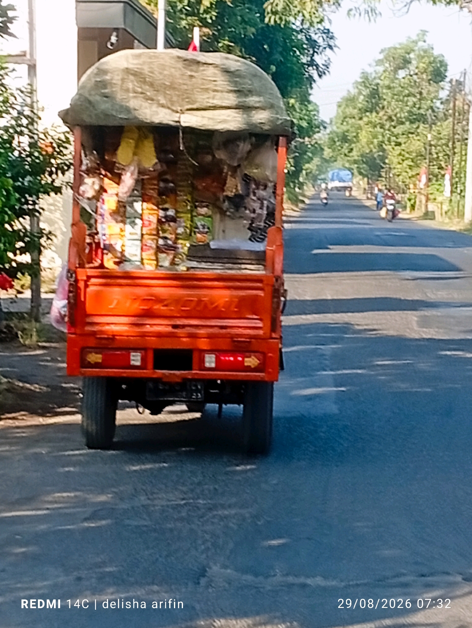 A rear view of an orange three-wheeled truck is captured on a paved road in Ngabar, Indonesia. The truck bed is filled with various products, some of which are packaged snacks in colorful wrappers, covered with a large piece of brown tarp. The truck is parked on the right side of the road. In the background, a few motorcycles and a blue truck are visible on the road, suggesting a rural or suburban setting with some traffic. Trees line both sides of the road, and the sky is bright, indicating daytime. The timestamp "29/08/2026 07:32" and "REDMI 14C | delisha arifin" are overlaid on the bottom of the image.