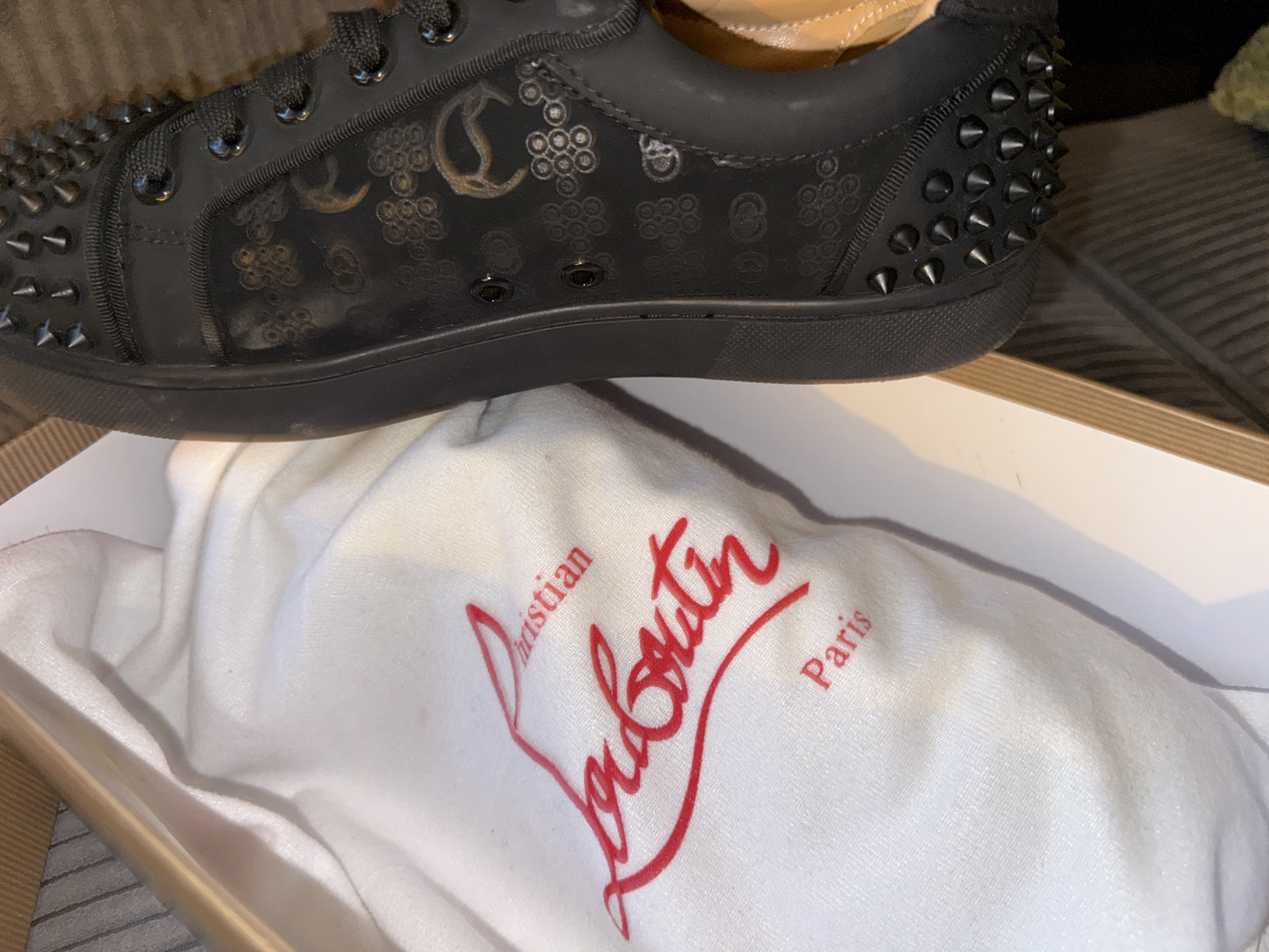 The image shows a close-up, angled view of a black Christian Louboutin sneaker with spike embellishments, placed inside an open shoebox with a white dust bag. The dust bag has the Christian Louboutin logo printed in red. The sneaker has a subtle patterned design embossed on its side, consisting of circular shapes and what appear to be stylized 'C' logos. The sole of the shoe is black, and the laces are also black. The interior of the shoe, visible at the top, is a tan leather. The shoebox is made of light-colored material, likely cardboard, and is resting on a textured surface, possibly a rug or striped fabric. The lighting suggests an indoor setting. There is no visible information within the image that would indicate the location being the city of Varna, Bulgaria.