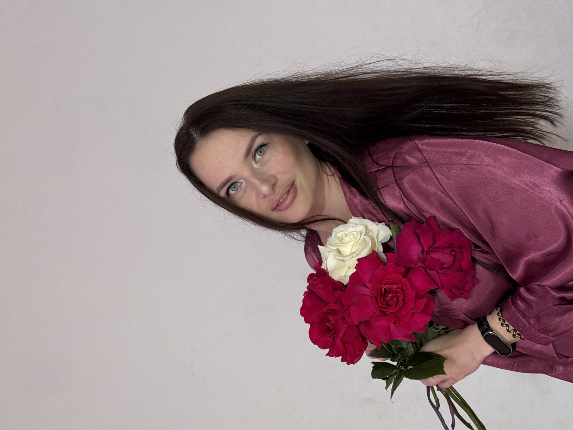 A medium shot captures a woman with long, dark brown hair and green eyes, holding a bouquet of red and white roses. She is wearing a silky, deep pink robe and a black smartwatch on her left wrist, accessorized with a coiled gold bracelet. The woman is positioned in front of a plain, light gray wall, suggesting an indoor setting. The image does not provide any visual information to determine the location as Rusalevka, Russia.