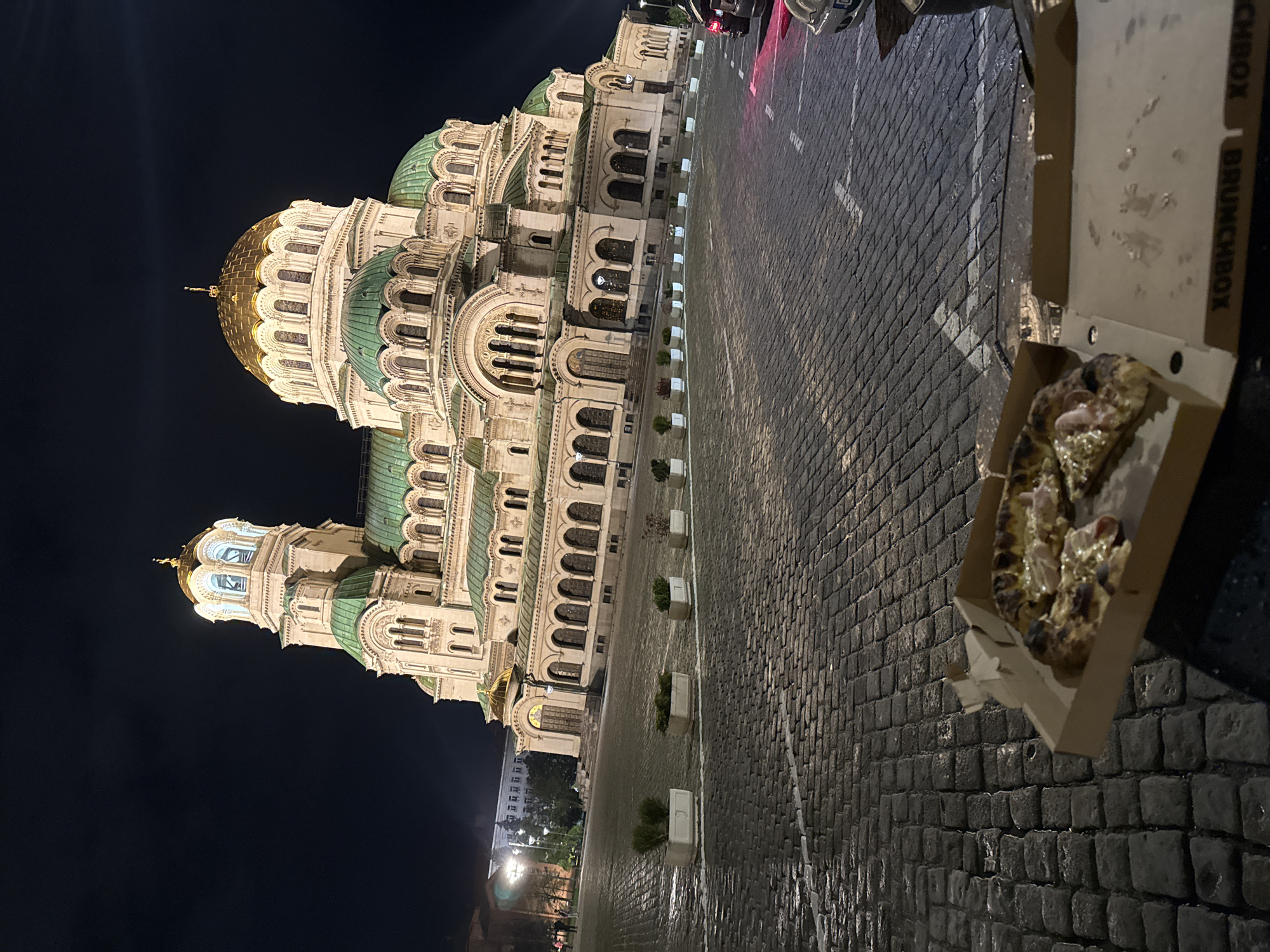 A nighttime shot shows the St. Alexander Nevsky Cathedral in Sofia, Bulgaria. The cathedral is brightly lit, contrasting with the dark sky. To the right is a cobblestone street with white lines marking lanes. An open pizza box with a partially eaten pizza sits on the ground next to the street. The box has “BRUNCHBOX” printed on its side. Cars are parked in the background near the cathedral. In the distance, across the street, are street lights illuminating a building. The ground appears slightly wet.