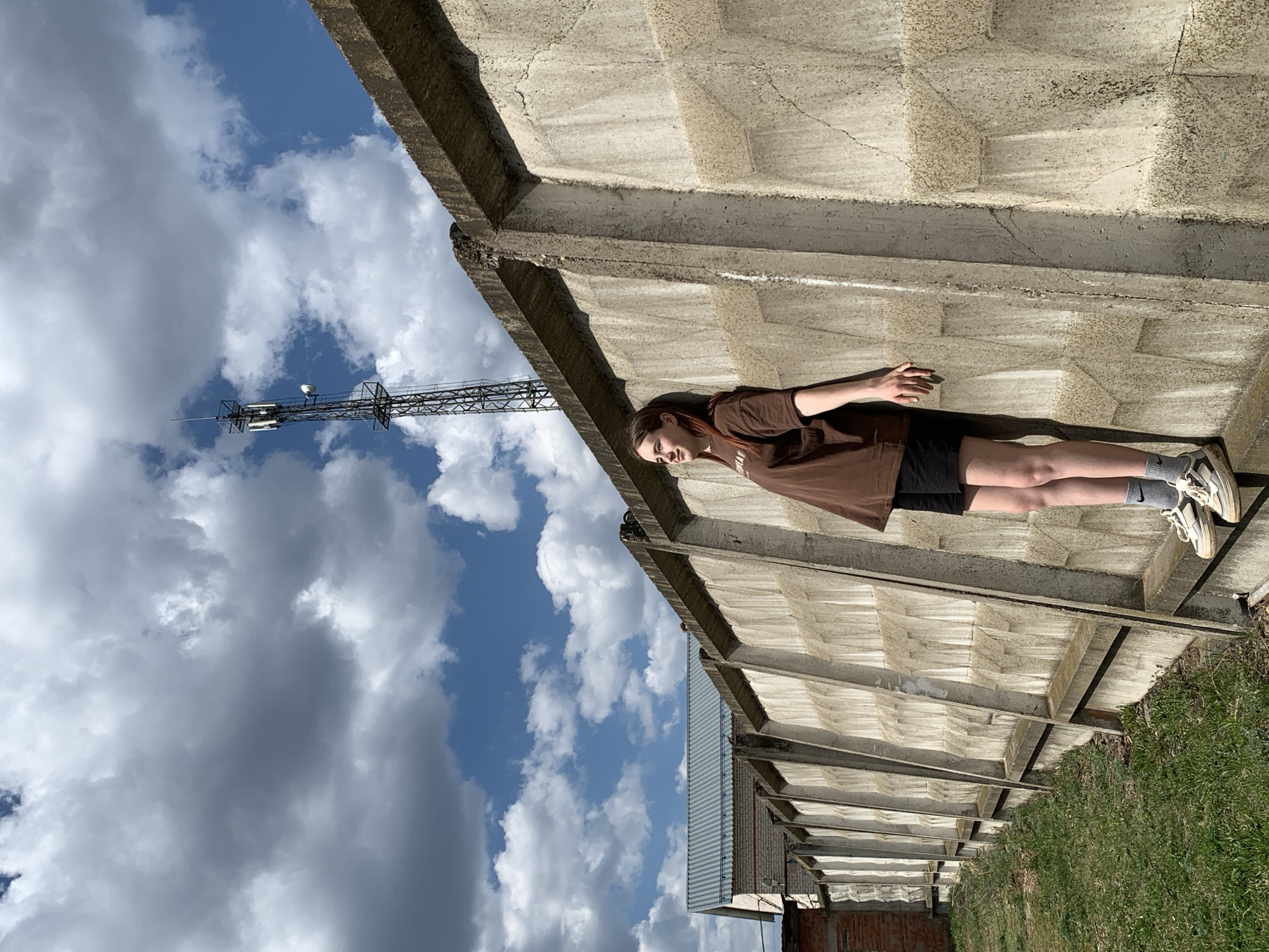A vertical medium shot reveals a young woman lying on a concrete structure, with a communication tower visible in the background, set against a sky with scattered clouds.
The woman is positioned in the center-right of the frame, reclining on what appears to be a portion of a concrete roof or elevated structure. She wears a brown short-sleeved shirt, black shorts, grey socks, and white sneakers.
The concrete structure has a distinct grid pattern, suggesting pre-fabricated panels. It is weathered, showing signs of age.
The background consists of a partly cloudy sky to the left, with a metal communication tower extending upwards. In the lower right, green grass and vegetation are visible, suggesting a rural or suburban setting. A brick building is also faintly visible.
Based on the information available, the location is identified as Фоки, Russia.