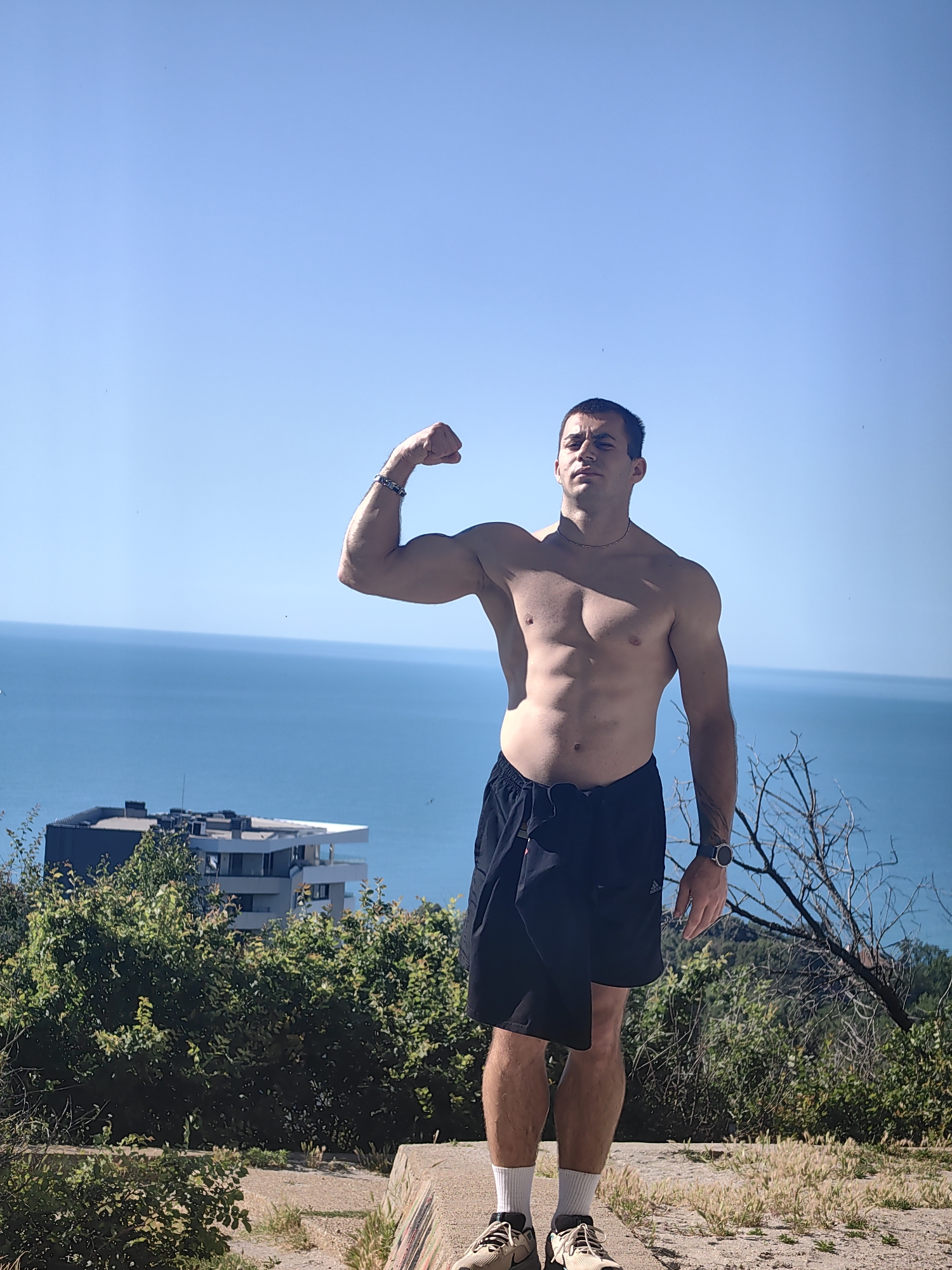 A shirtless man with athletic build stands outdoors, flexing his bicep. He wears black shorts, white socks, and athletic shoes. A watch is visible on his left wrist. Behind him, a body of water, likely the sea, stretches to the horizon under a clear blue sky. In the middle ground, a modern building with multiple levels is nestled amongst lush green foliage. The man is positioned on a raised concrete surface, with dry grass and a bare tree branch in the foreground. The lighting suggests it is daytime.