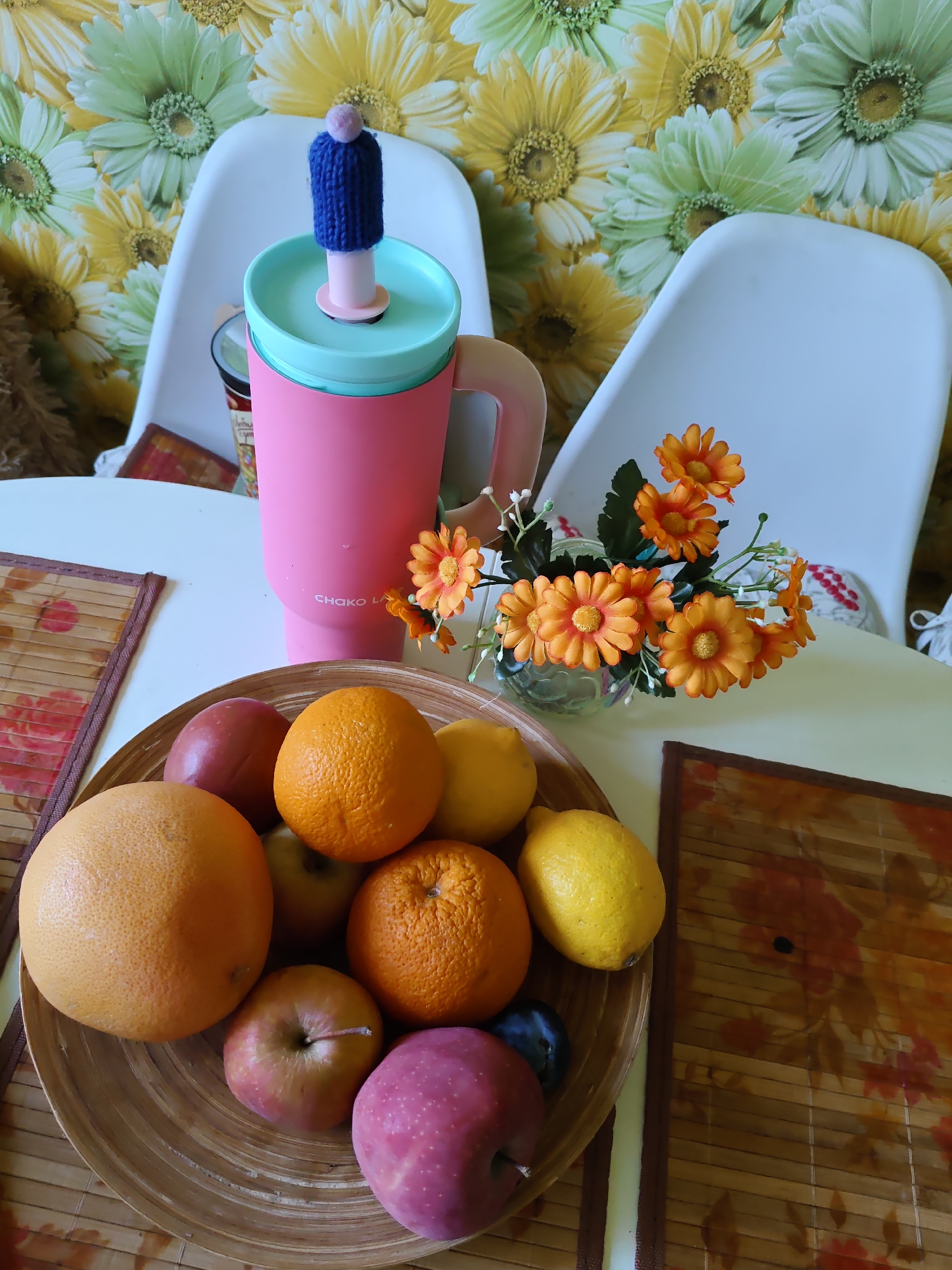 The image depicts a still life arrangement on a white table, set against a floral-patterned wallpaper. In the foreground, a wooden bowl overflows with various fruits, including oranges, apples, and lemons. To the right of the bowl, a small bouquet of artificial orange flowers in a glass vase adds a touch of color. Behind the fruit bowl, a pink and teal travel mug with a knitted blue topper and a pink pom-pom stands prominently. Two white chairs are visible in the background, with a patterned placemat on the left side of the table and another on the right, partially obscured by the fruit. The overall scene suggests a domestic setting. The image was taken in Rostov-on-Don, Russia.